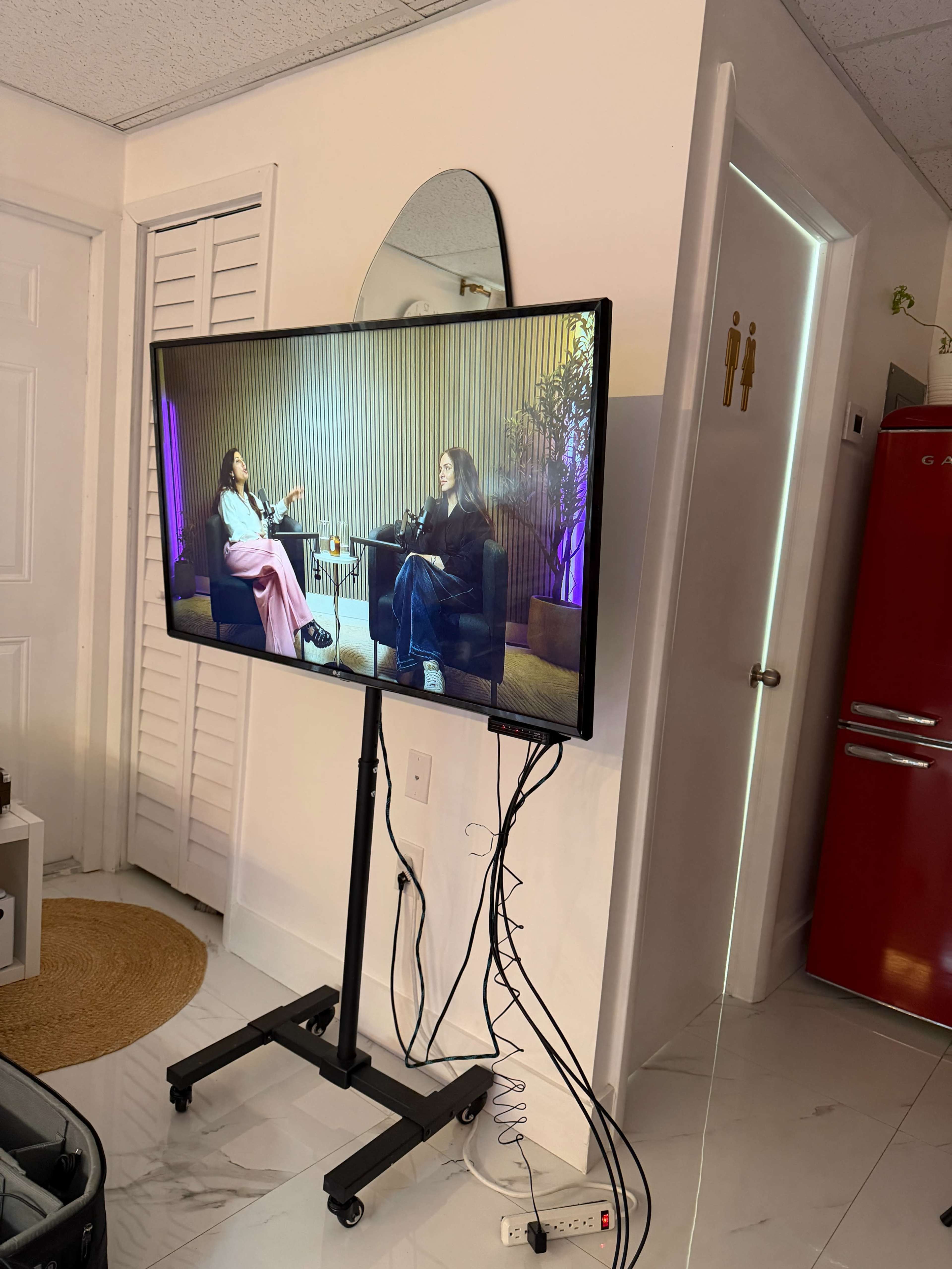 A television on a stand displays two women seated and conversing in a brightly lit room with modern decor.