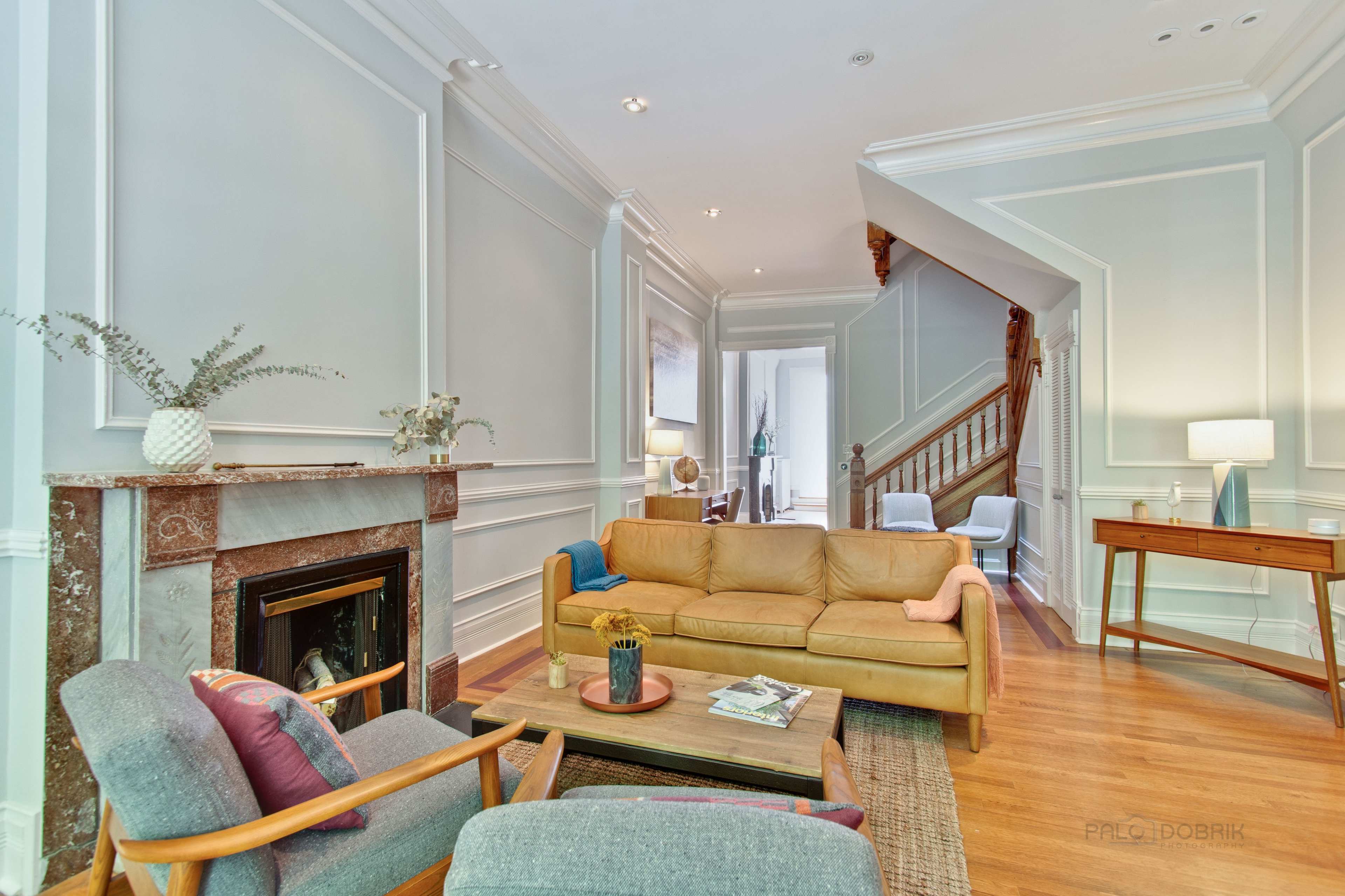 A living room with a tan leather sofa, a wooden coffee table, and a fireplace, adjacent to a staircase and a well-lit hallway.