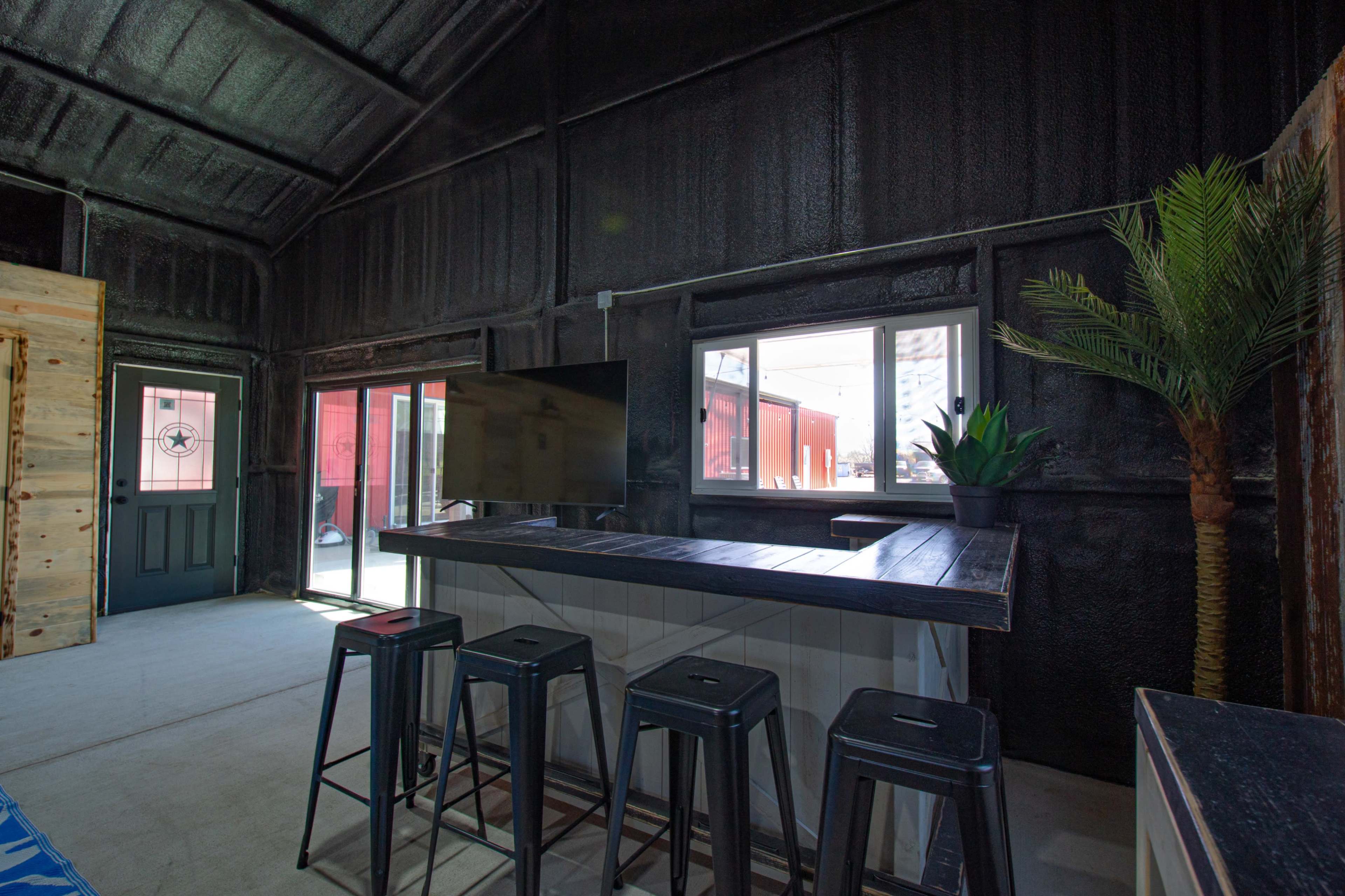 The image shows a modern interior space featuring a bar-style table with black stools, a television mounted on the wall, and large windows that provide natural light.