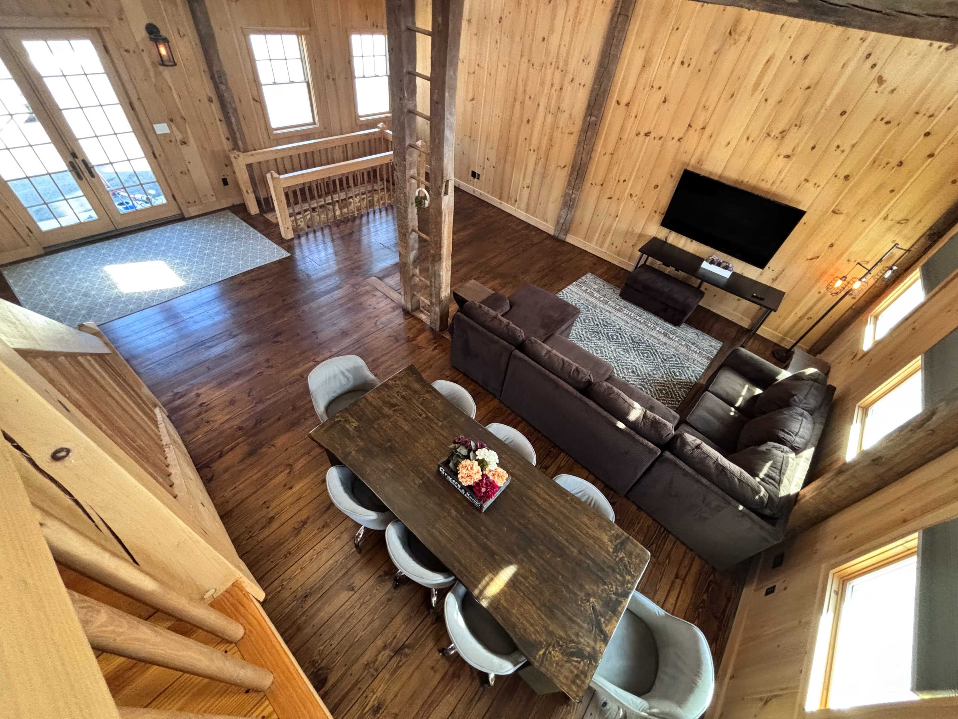 The image shows a wooden interior of a cozy living space featuring a dining table with chairs, a sofa set, and a television, illuminated by natural light from large windows.