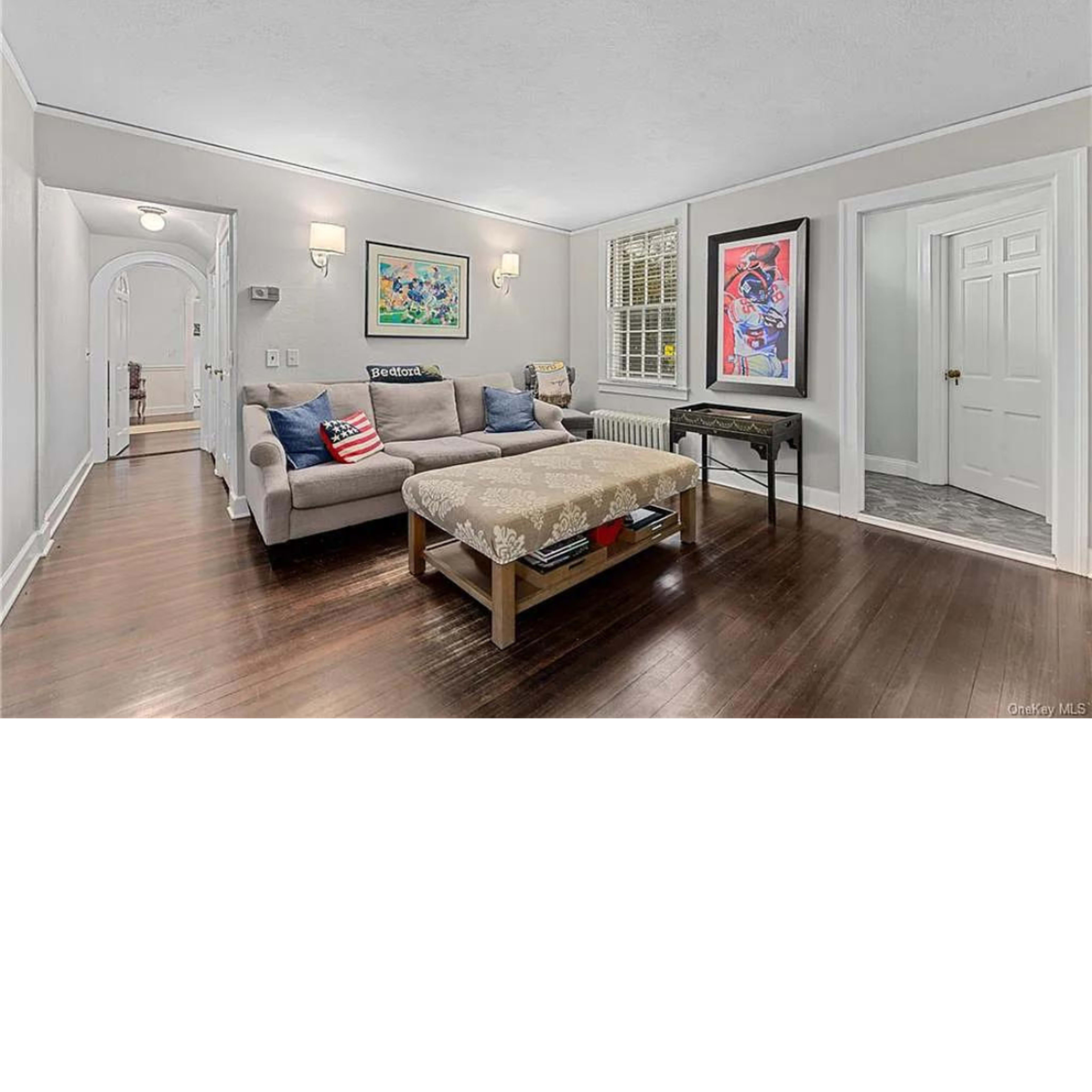 A neutral living room features a sofa with decorative pillows, a coffee table, and artwork on the walls, with a doorway leading to a hallway.