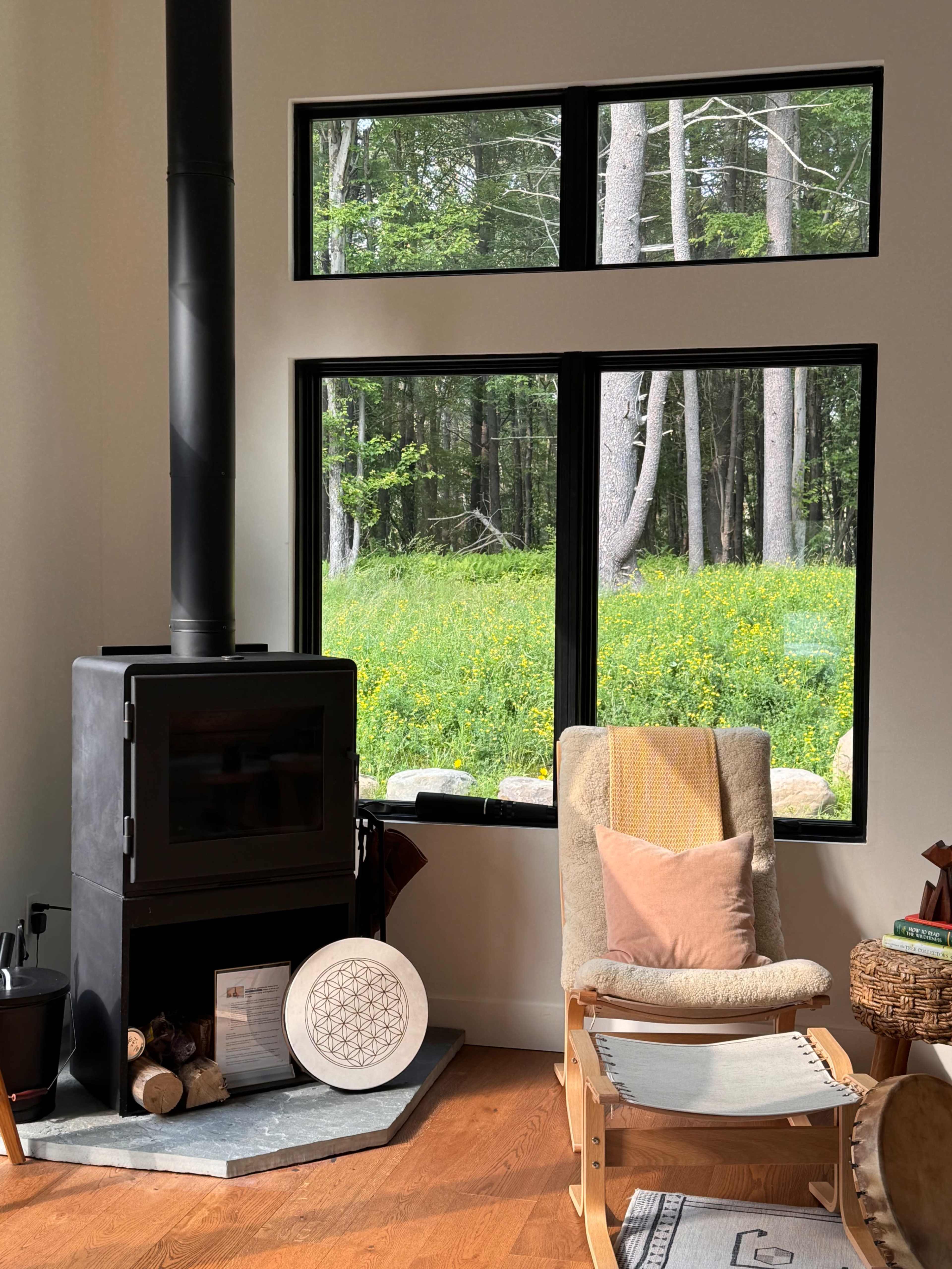 A wood stove stands in the corner of a room, next to a rocking chair, with large windows framing a view of a lush green landscape.