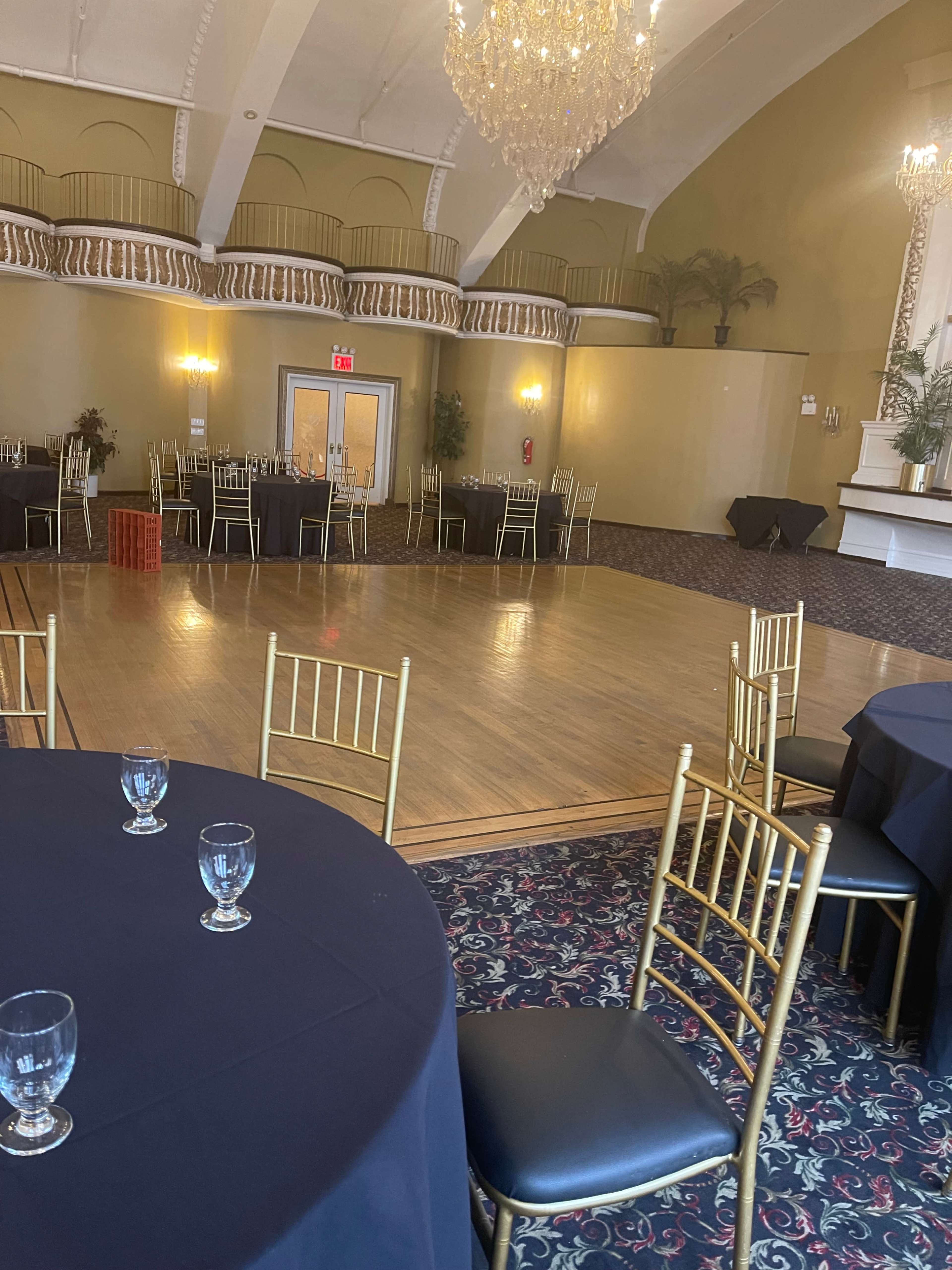 The image shows an elegant banquet hall with wooden floors, round tables covered in black cloth, and gold chairs arranged around the space, featuring a stage and a chandelier overhead.