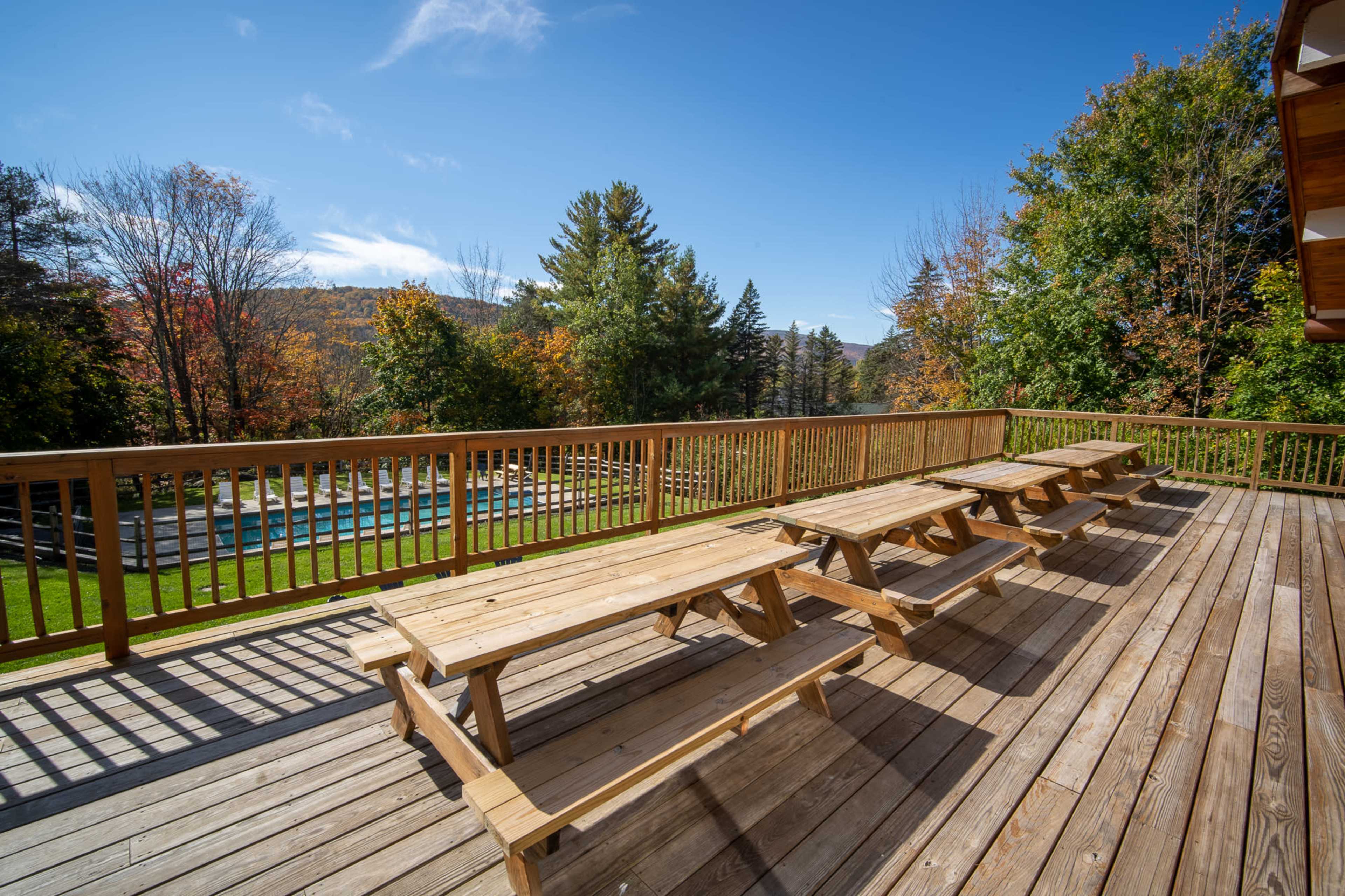 A wooden deck with multiple picnic tables overlooks a green lawn and a swimming pool surrounded by trees.