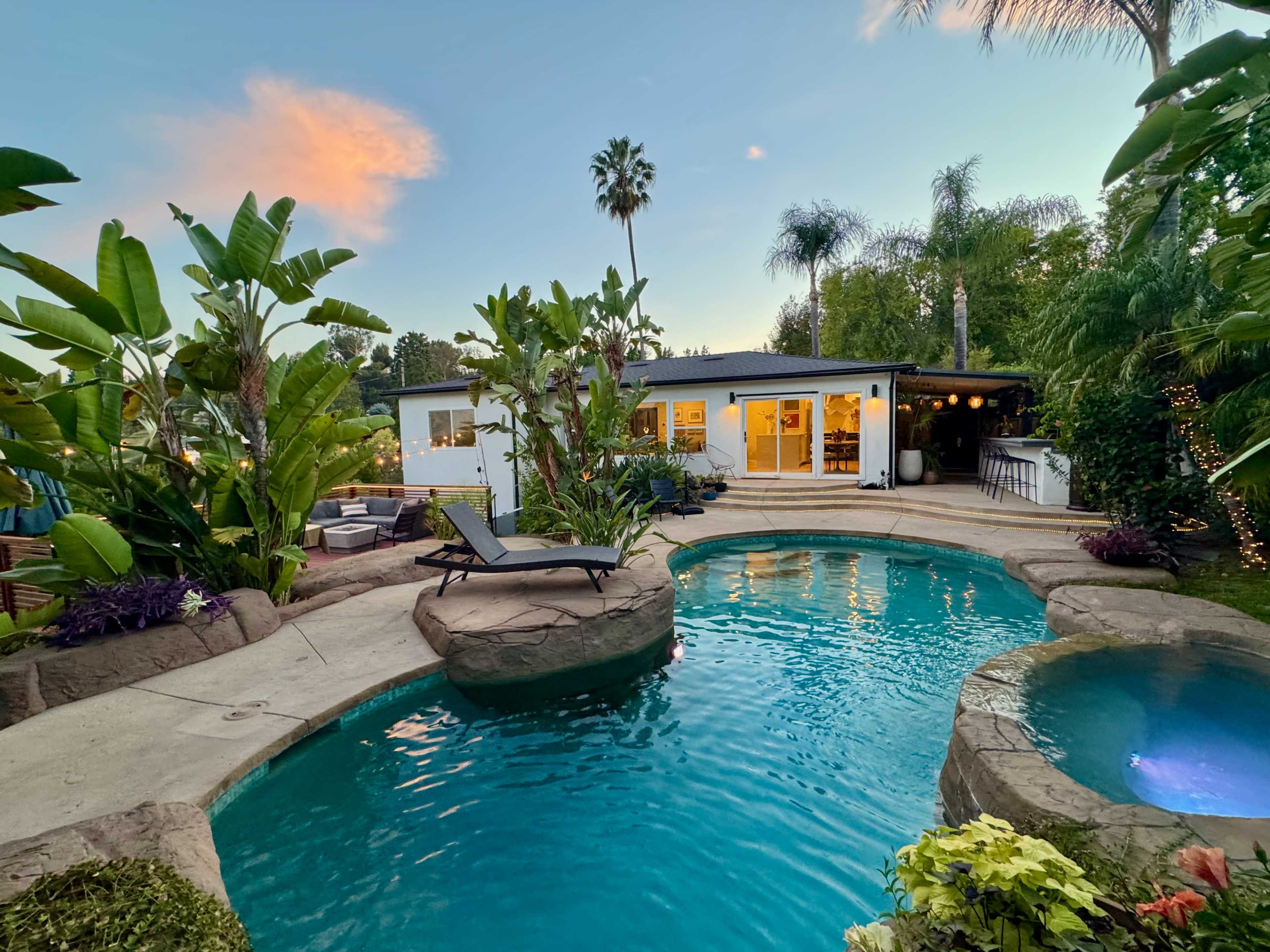The image shows a backyard with a swimming pool, a lounge area with seating, and a house in the background surrounded by palm trees and lush greenery.