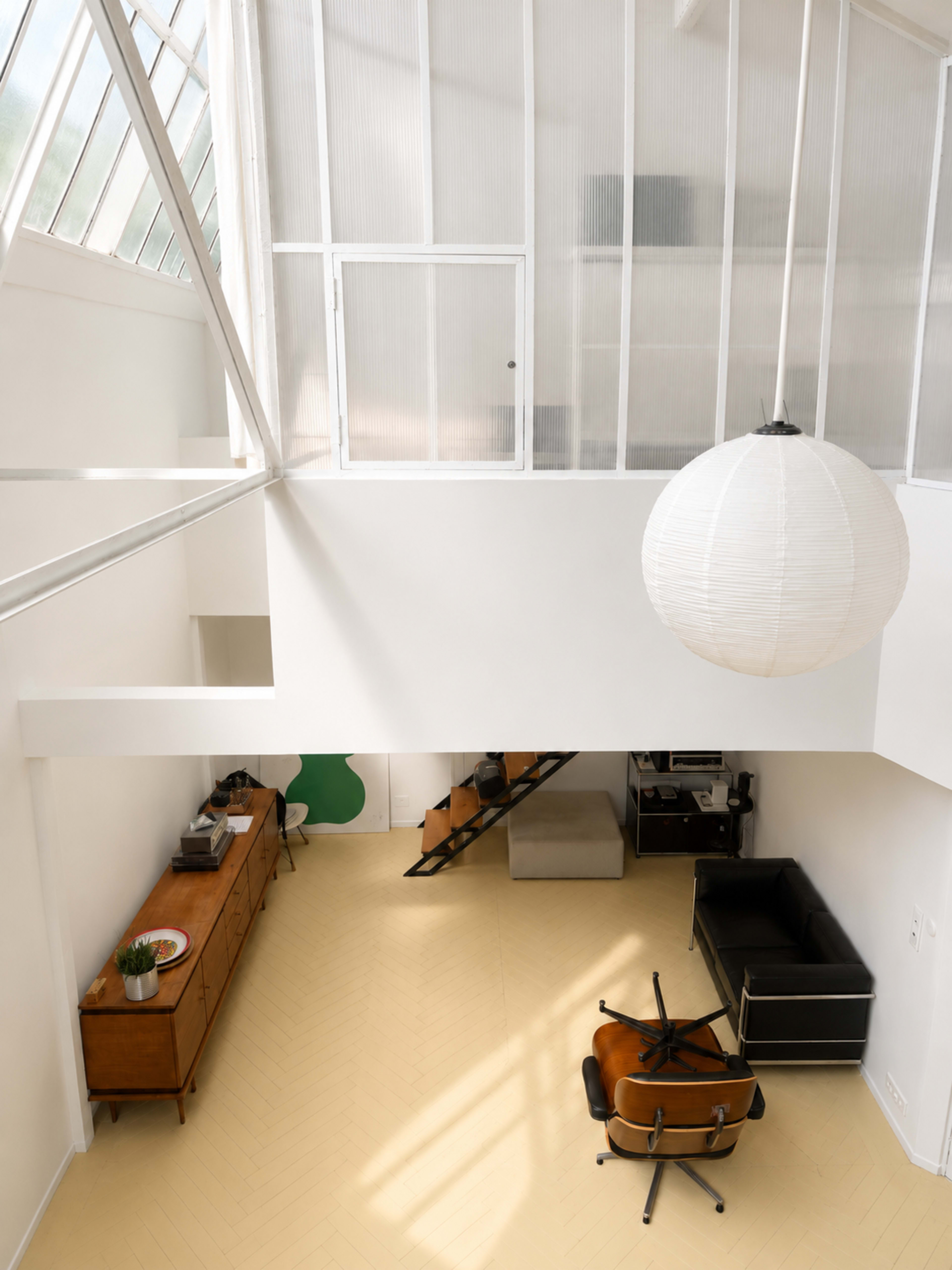 The image shows a spacious, modern interior with high ceilings, a light-colored floor, a black couch, a wooden dresser, and a hanging paper lantern.