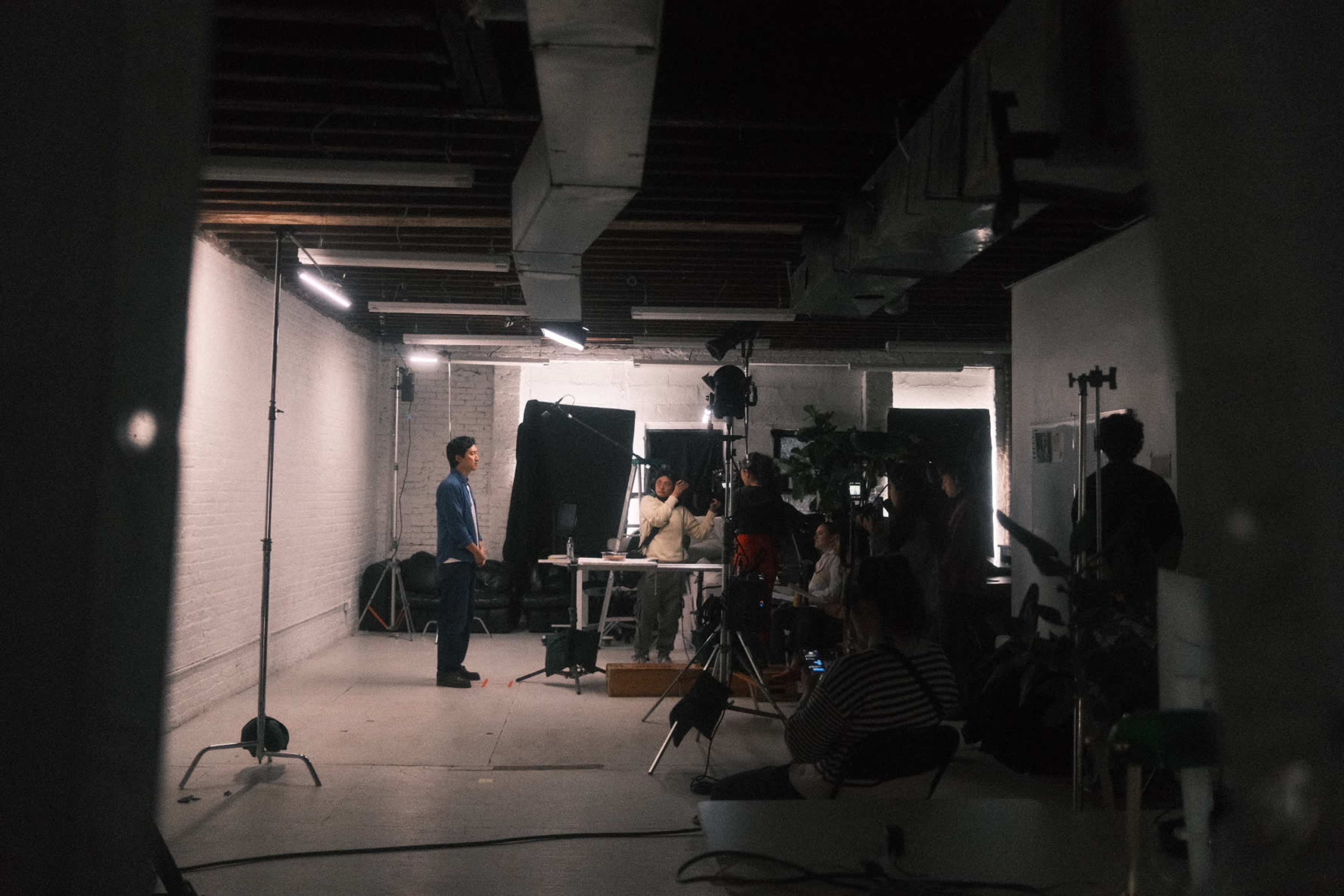 A man stands in front of a camera and lights while a group of people observes in a studio setting.