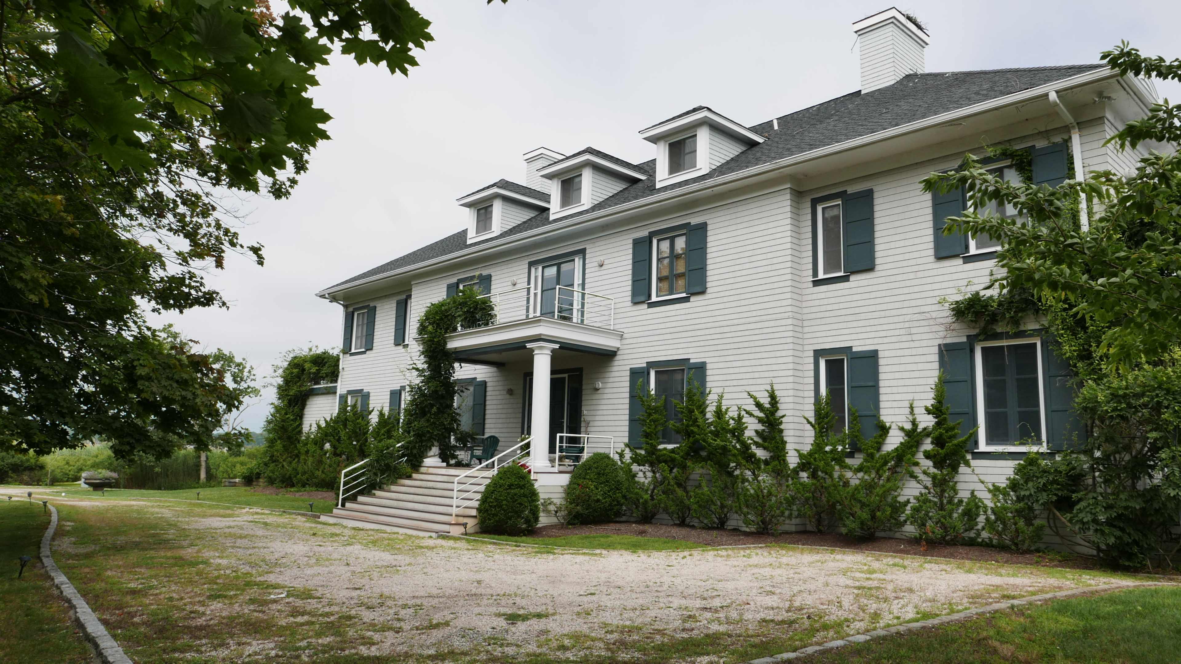 A large, two-story white house with multiple windows and a landscaped front yard sits along a gravel pathway.