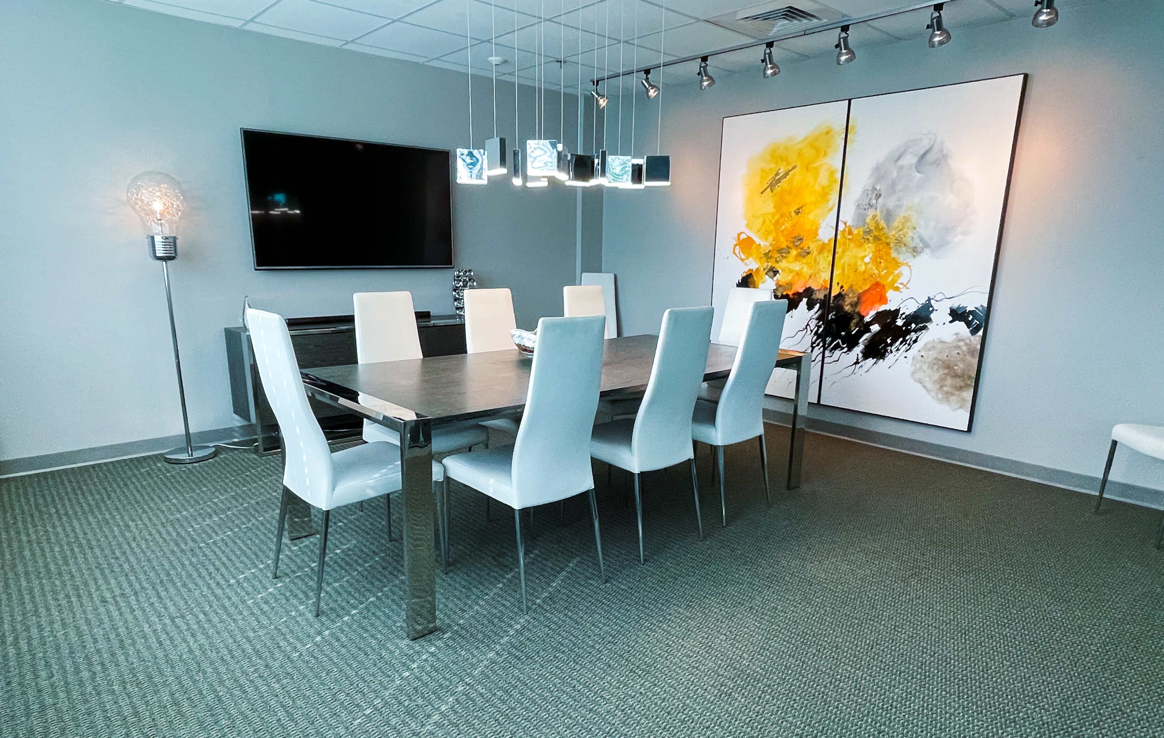 A modern conference room with a long table surrounded by white chairs, a large artwork on the wall, and a television mounted above a side cabinet.