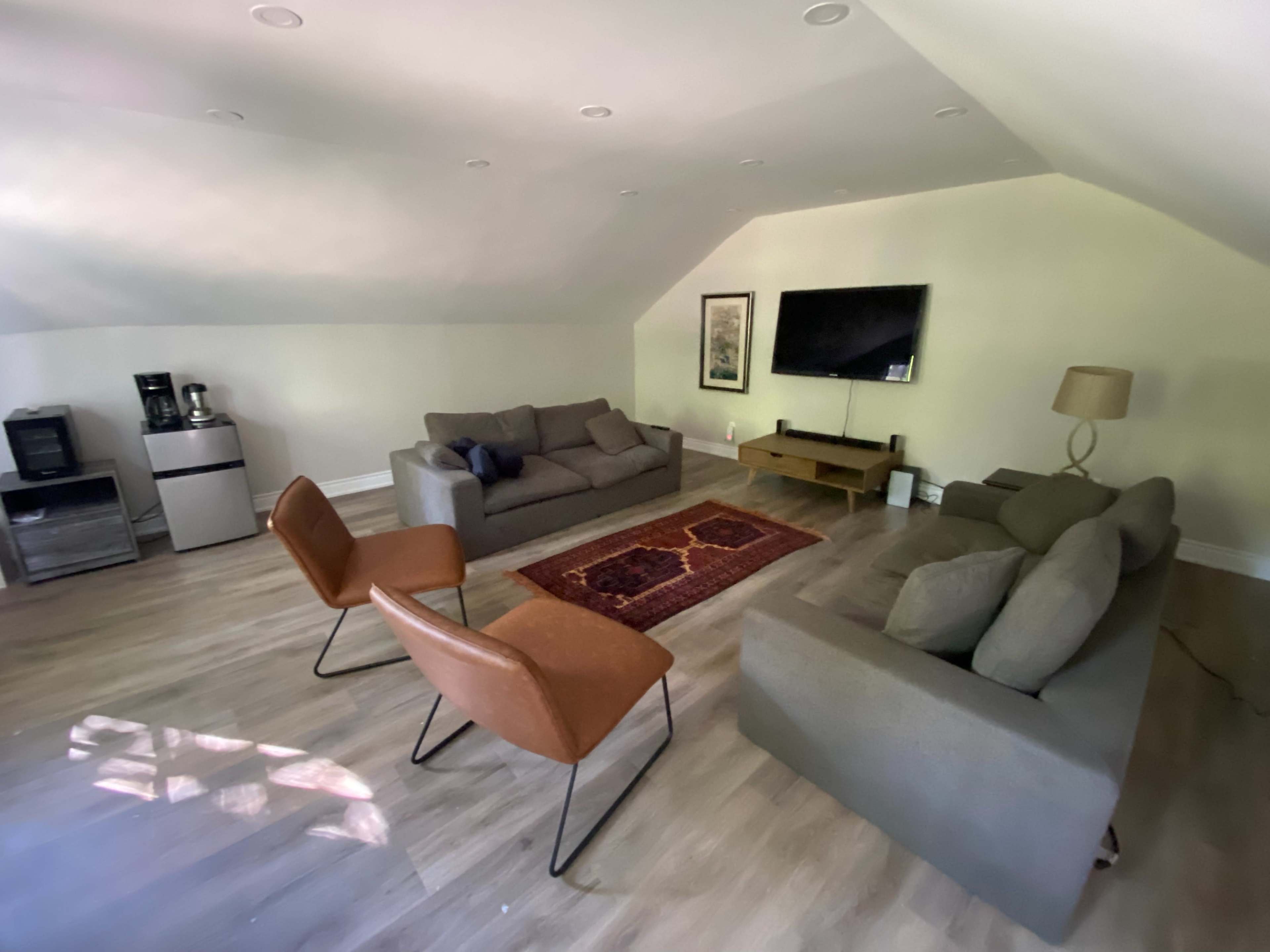 The image shows a spacious living area with two sofas, two chairs, a coffee table, a television mounted on the wall, and a kitchenette in the corner.