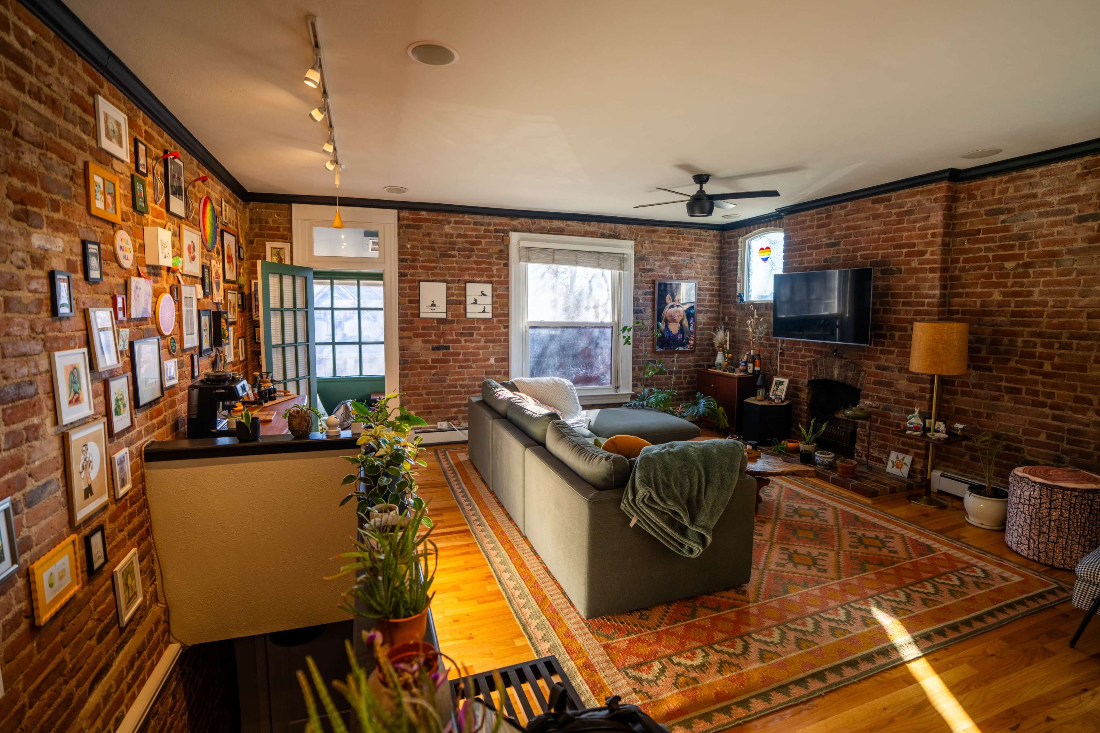 The image shows a cozy living room with exposed brick walls, a green couch, a coffee table, and a gallery wall filled with framed pictures.