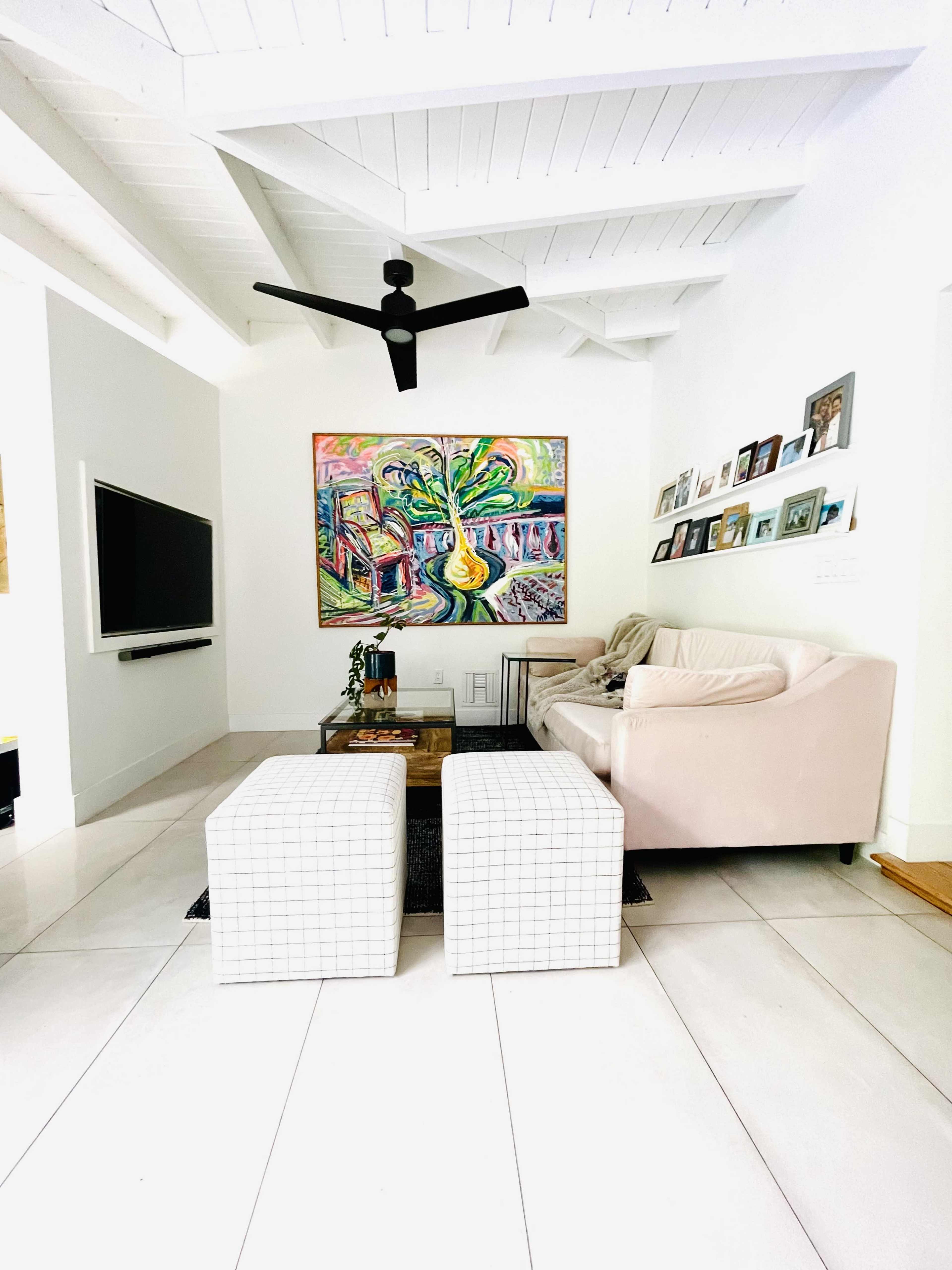 A modern living room features a pink couch, two square white ottomans, and a large colorful painting on the wall, with a television mounted opposite.