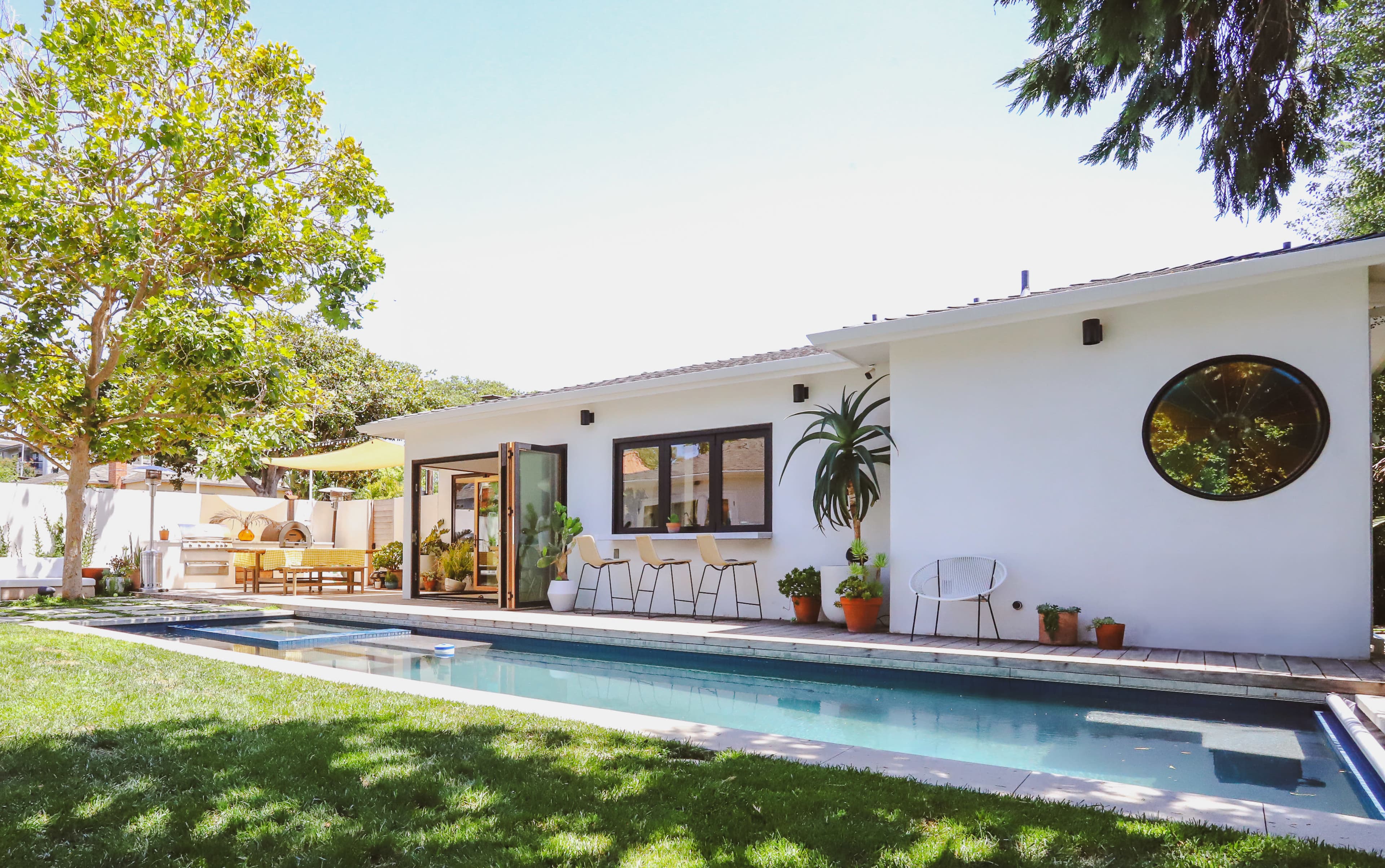 A modern single-story house with large windows overlooks a rectangular swimming pool and a landscaped backyard featuring trees and potted plants.