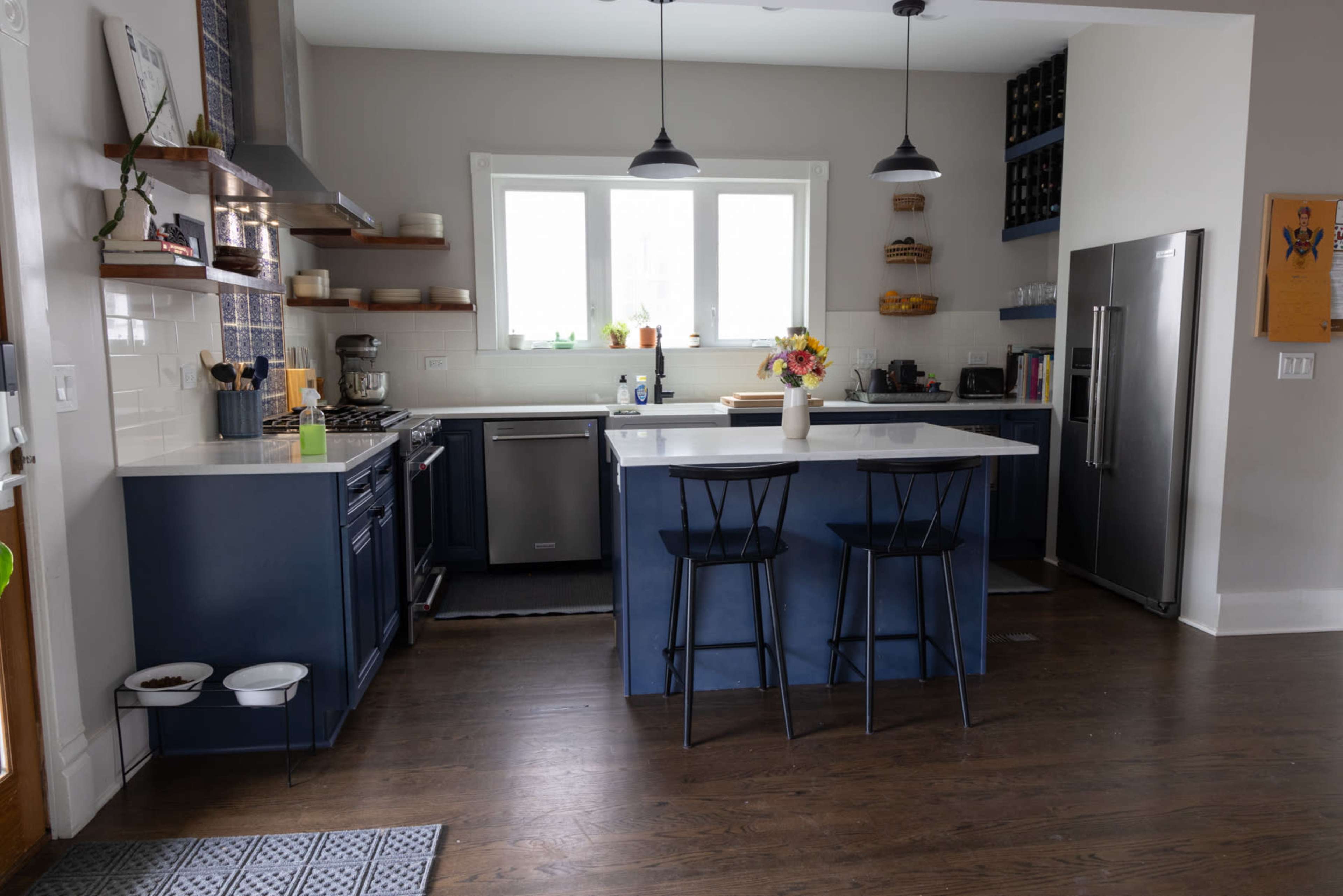 A modern kitchen with blue cabinetry, a large island with black stools, and stainless steel appliances.