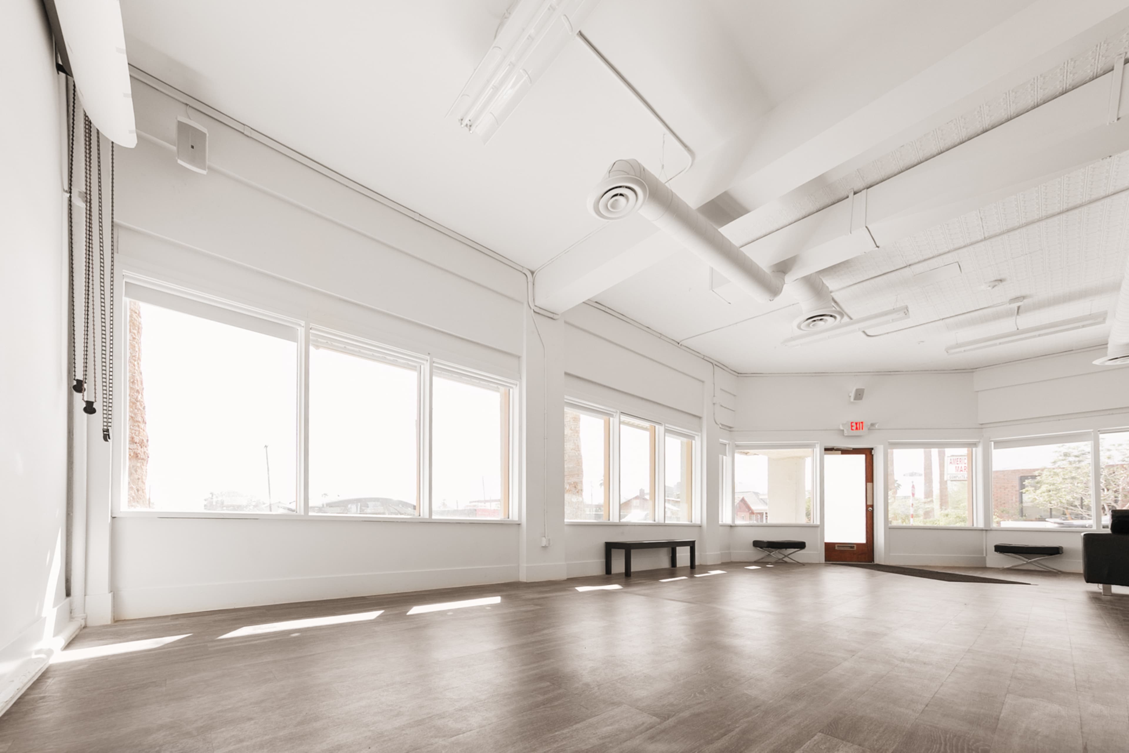 A spacious, brightly lit room with large windows and minimal furnishings, featuring a wooden floor and a single bench against the wall.