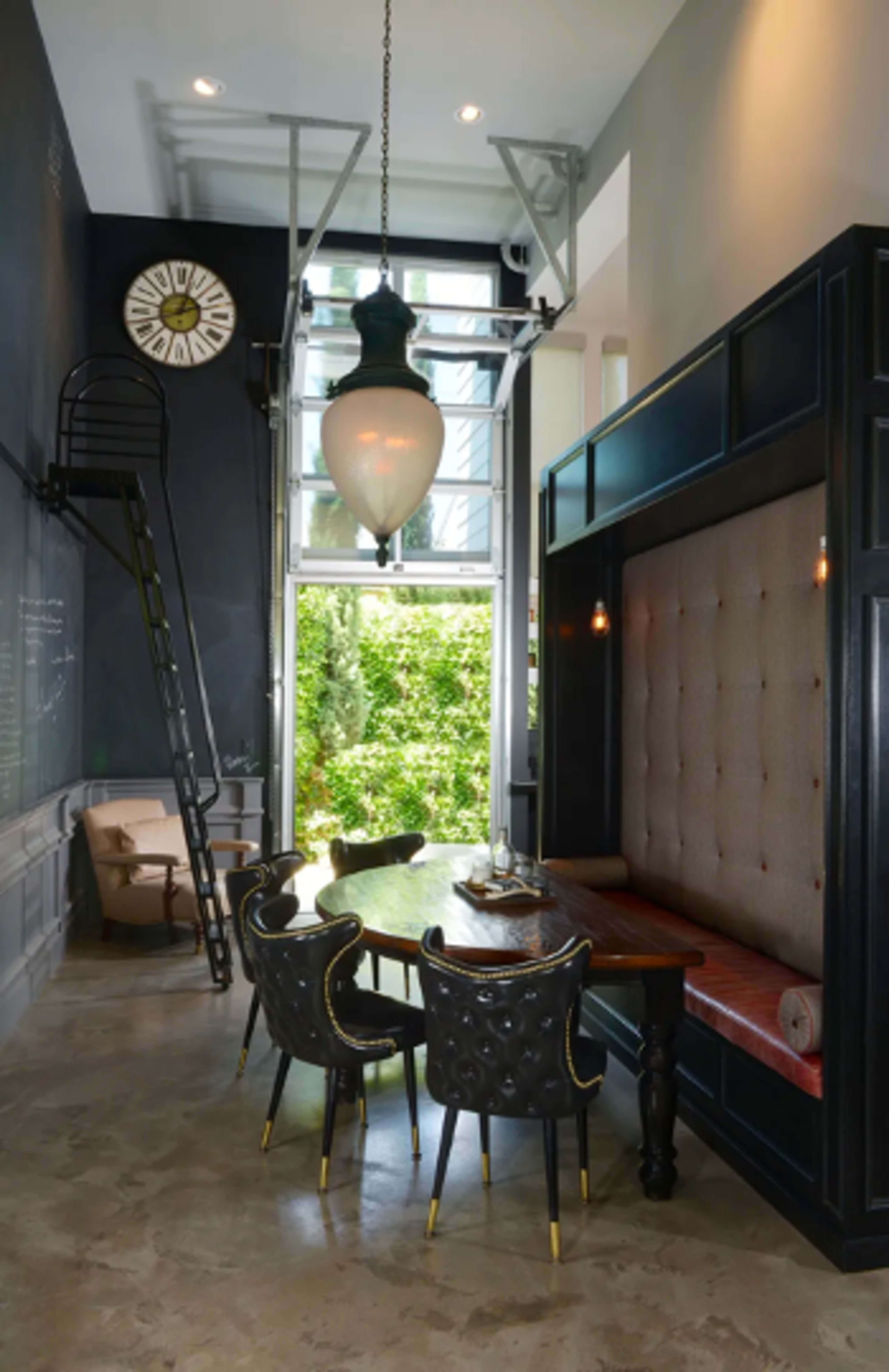 A modern dining area features a large wooden table surrounded by elegant chairs, with a plush seating nook and a tall staircase leading to a higher level.