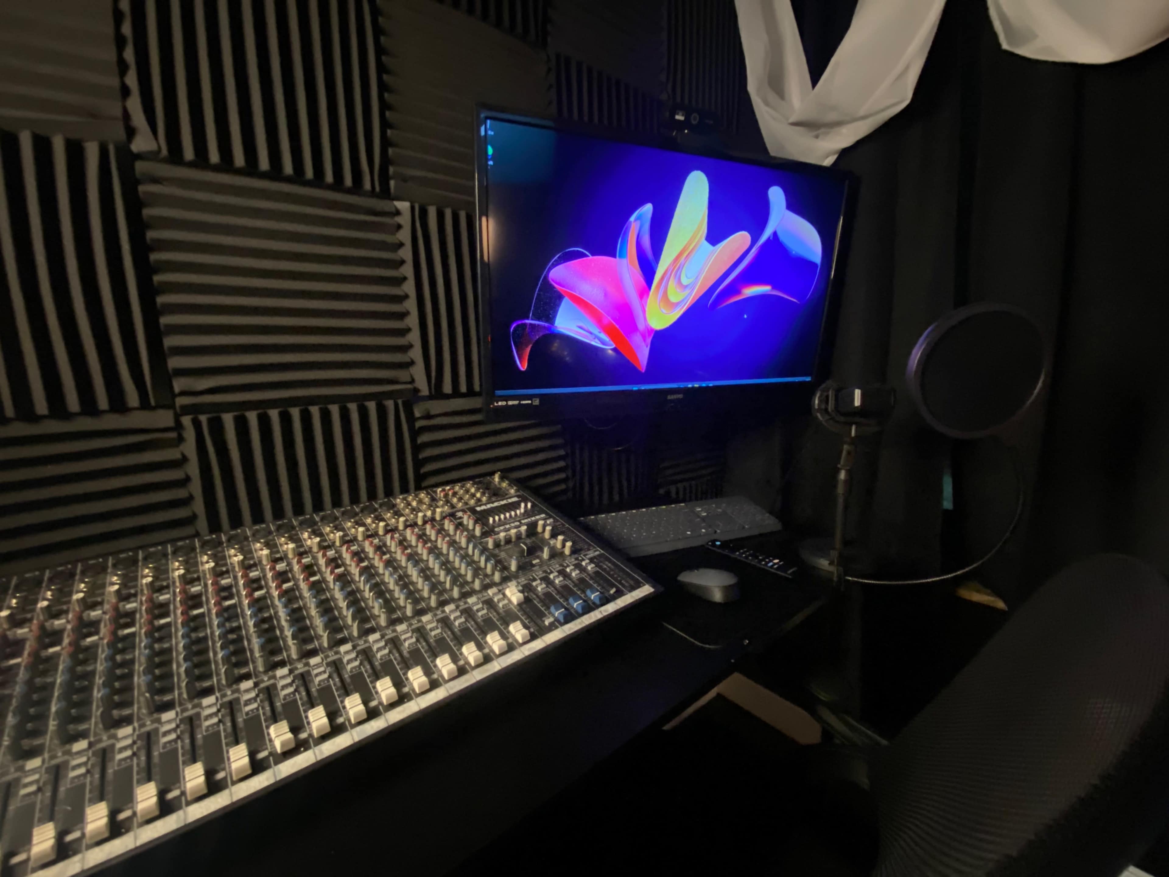 The image shows a recording studio setup with a large audio mixing console, a computer monitor displaying colorful graphics, and a microphone on a stand.