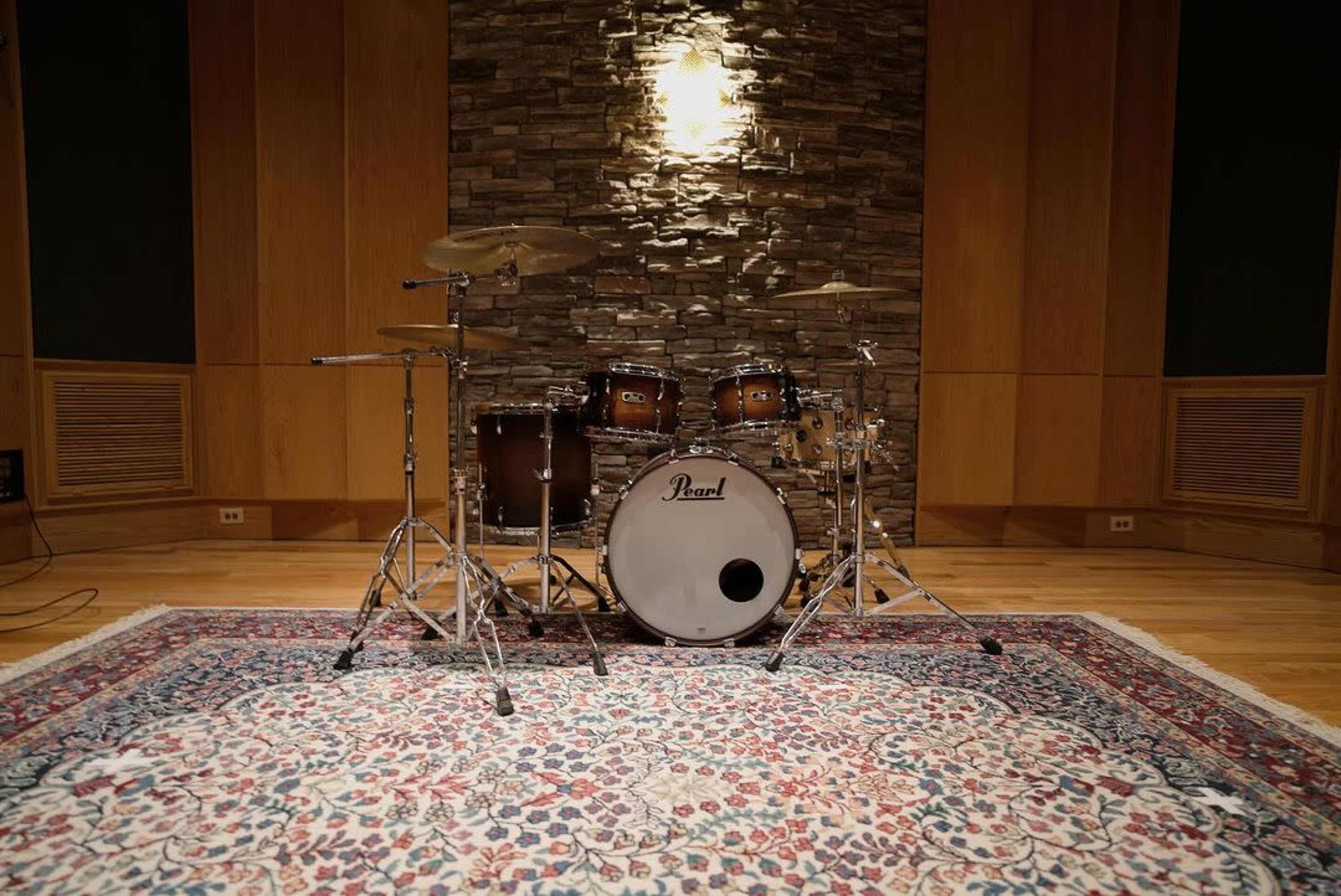 A drum set is positioned on a patterned rug in a studio with wooden walls and a stone backdrop.