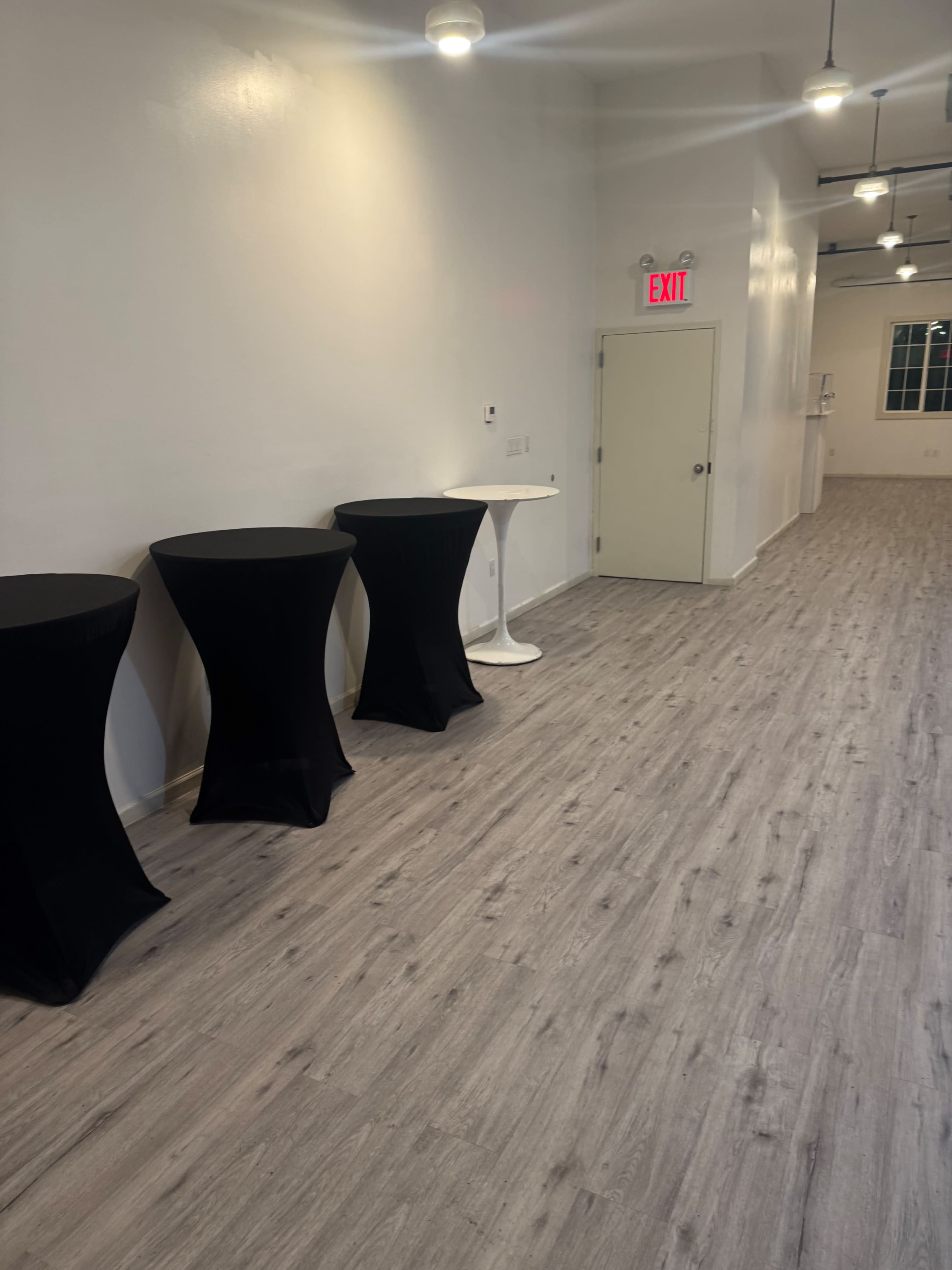 The image shows a narrow, well-lit hallway with three tall, round tables covered in black and white tablecloths along one side.