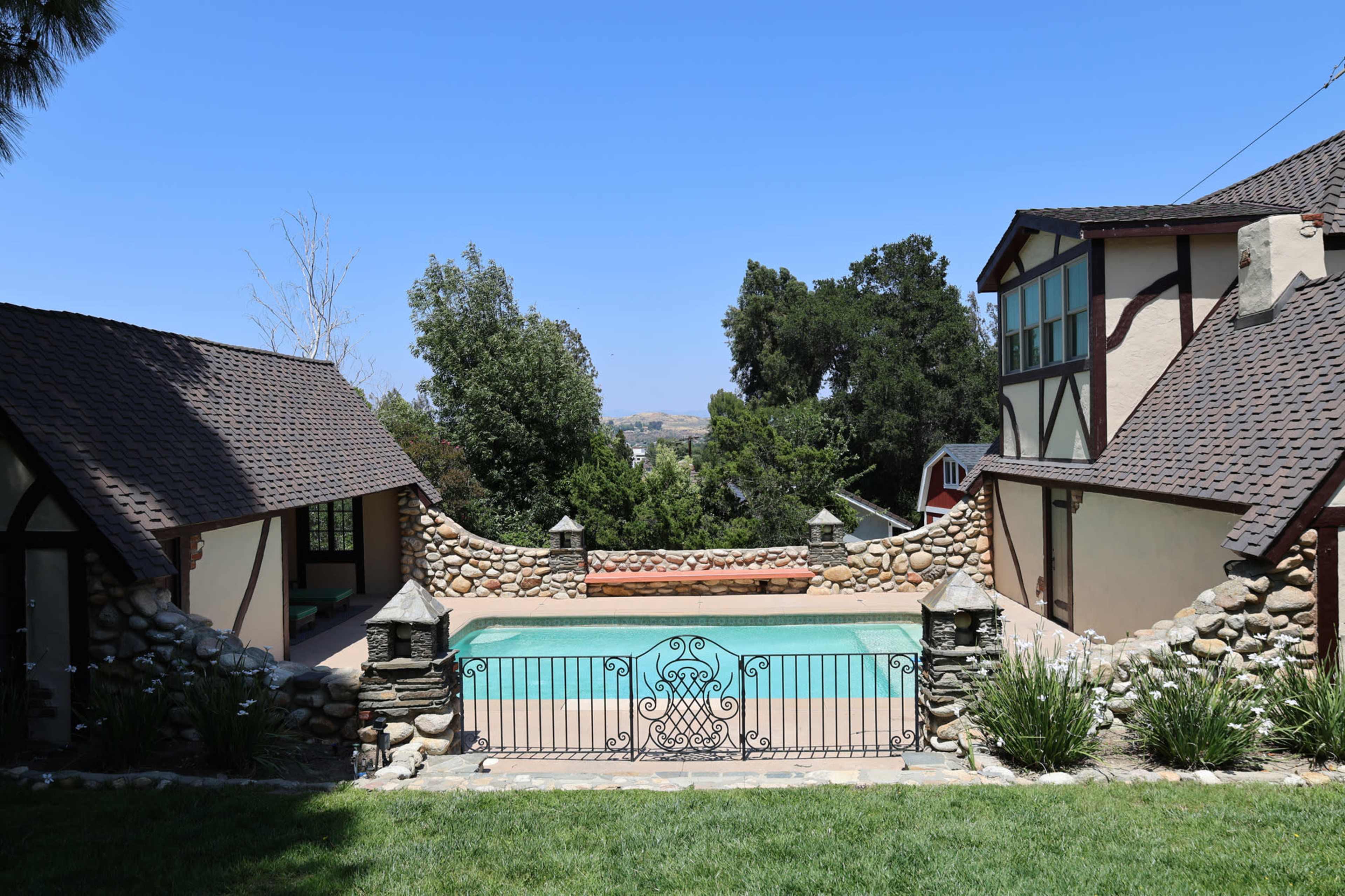 The image shows a stone-walled backyard with a rectangular swimming pool surrounded by greenery and two adjacent buildings.