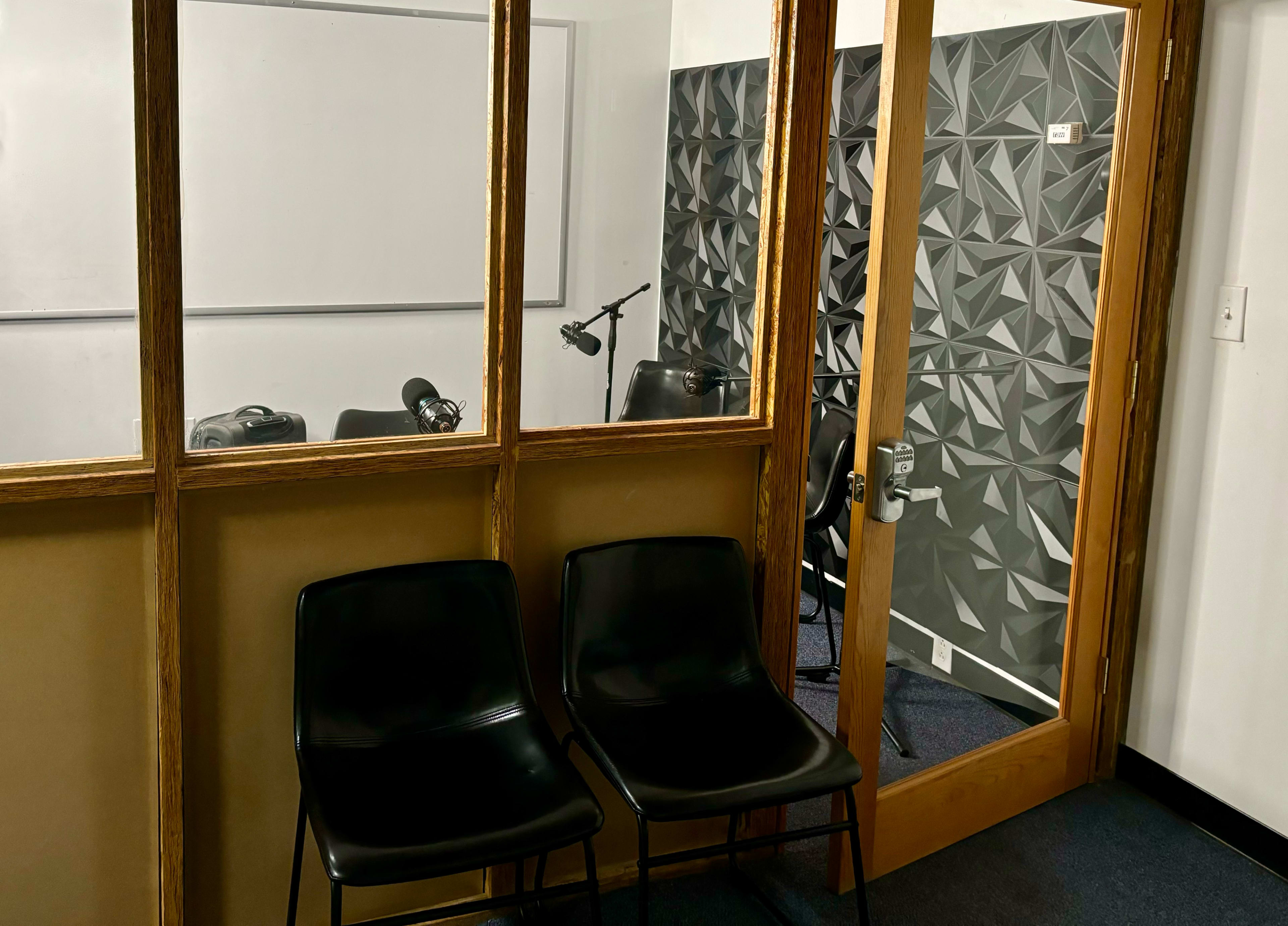 The image shows a small recording or interview room with two black chairs in the foreground and a textured wall with a microphone setup visible through a glass partition.