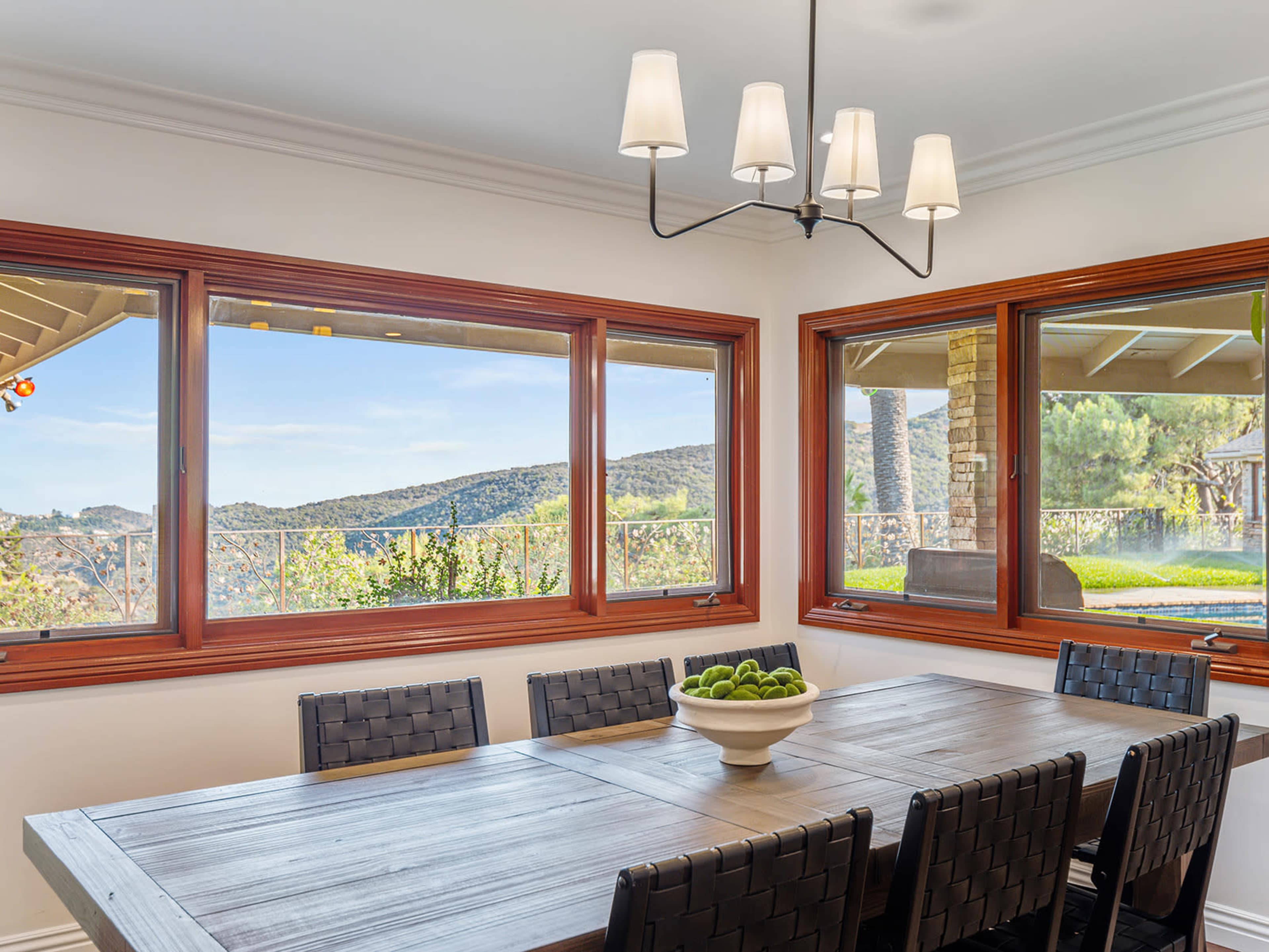 A dining room features a large wooden table surrounded by black woven chairs, with large windows showcasing a scenic view of green hills.