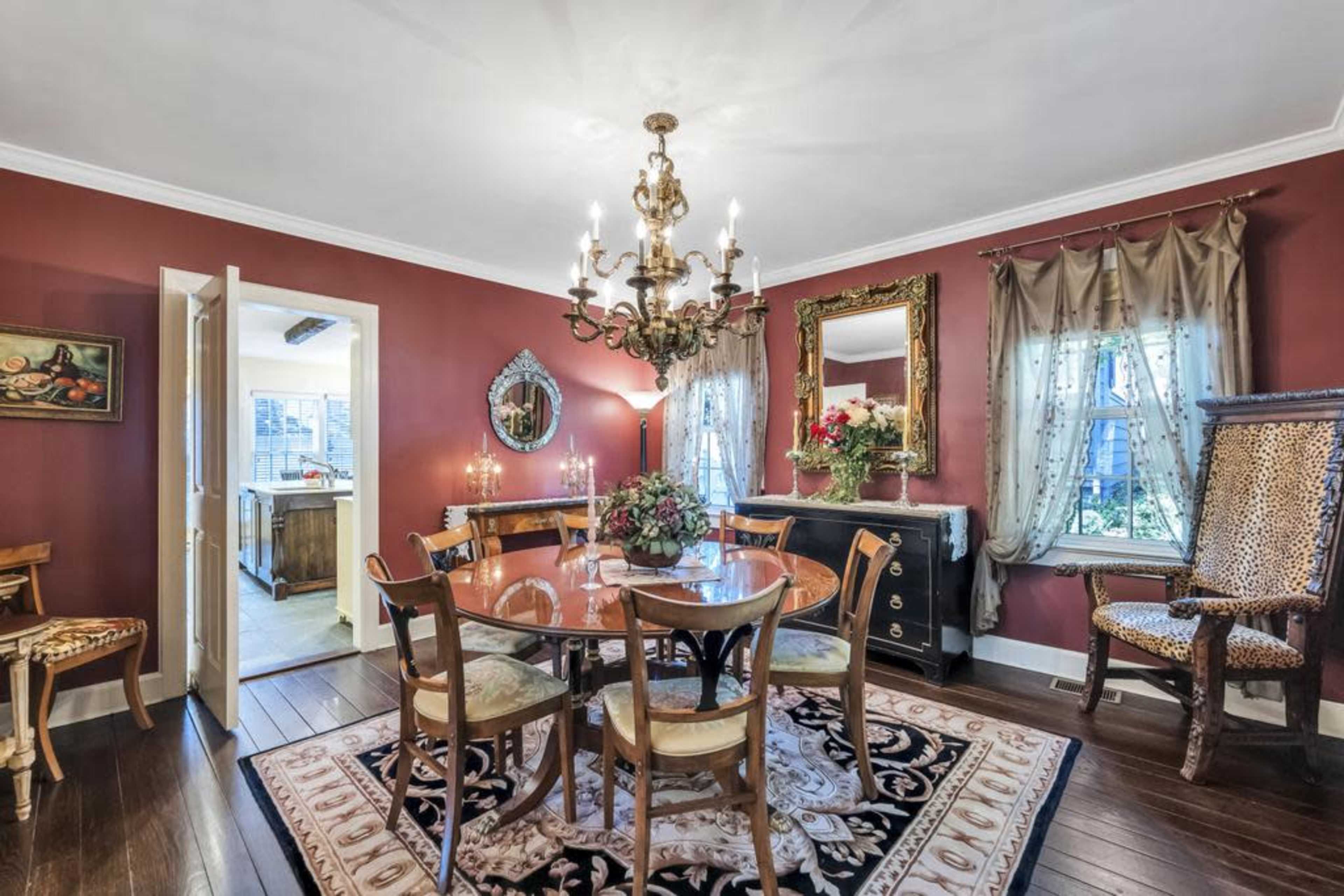 A dining room featuring a round table with chairs, a chandelier overhead, and red walls adorned with decor and large windows.