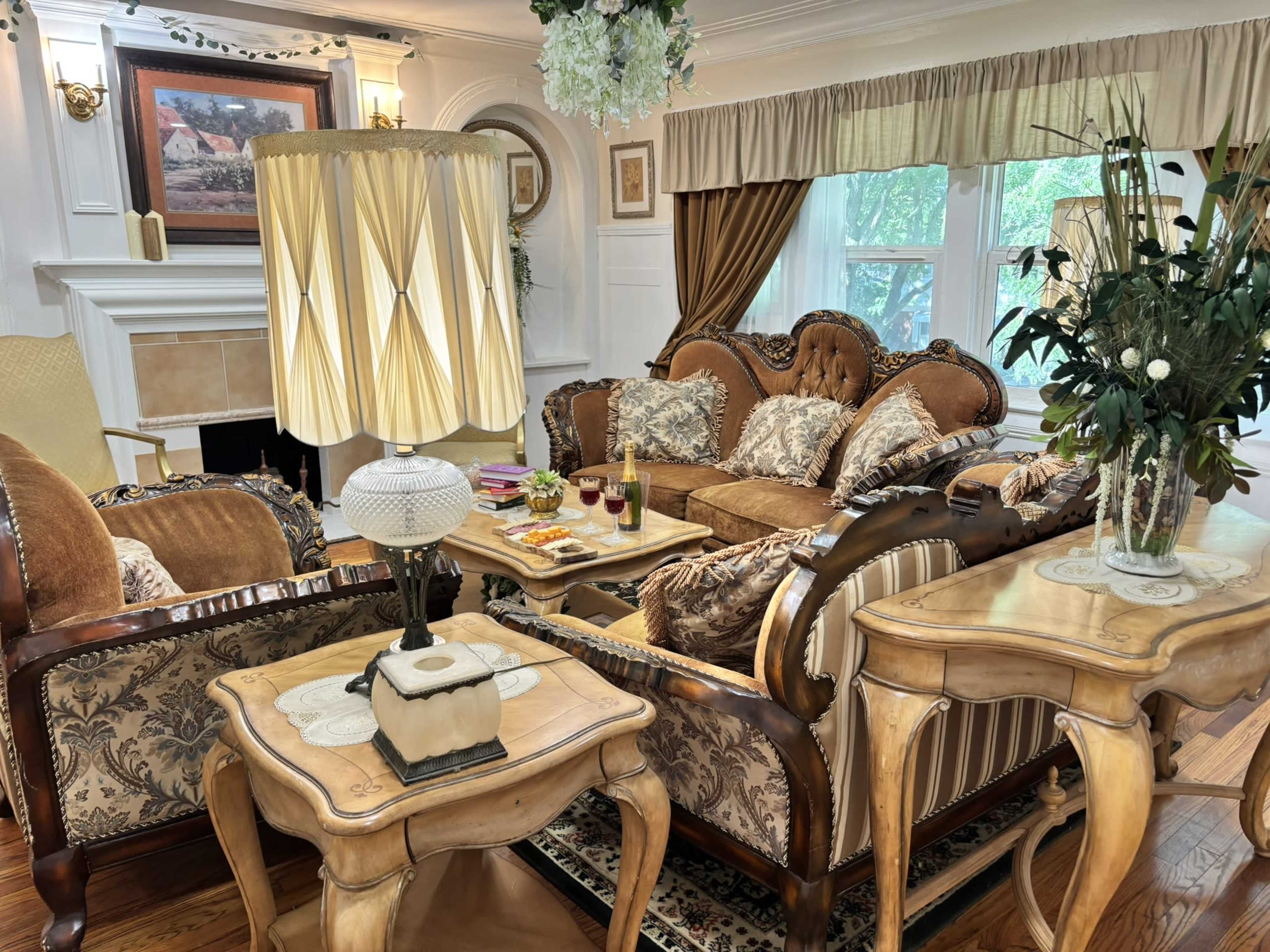 The image shows a elegantly furnished living room with a mix of ornate furniture, a coffee table set with drinks, and a decorative lamp beside the seating area.