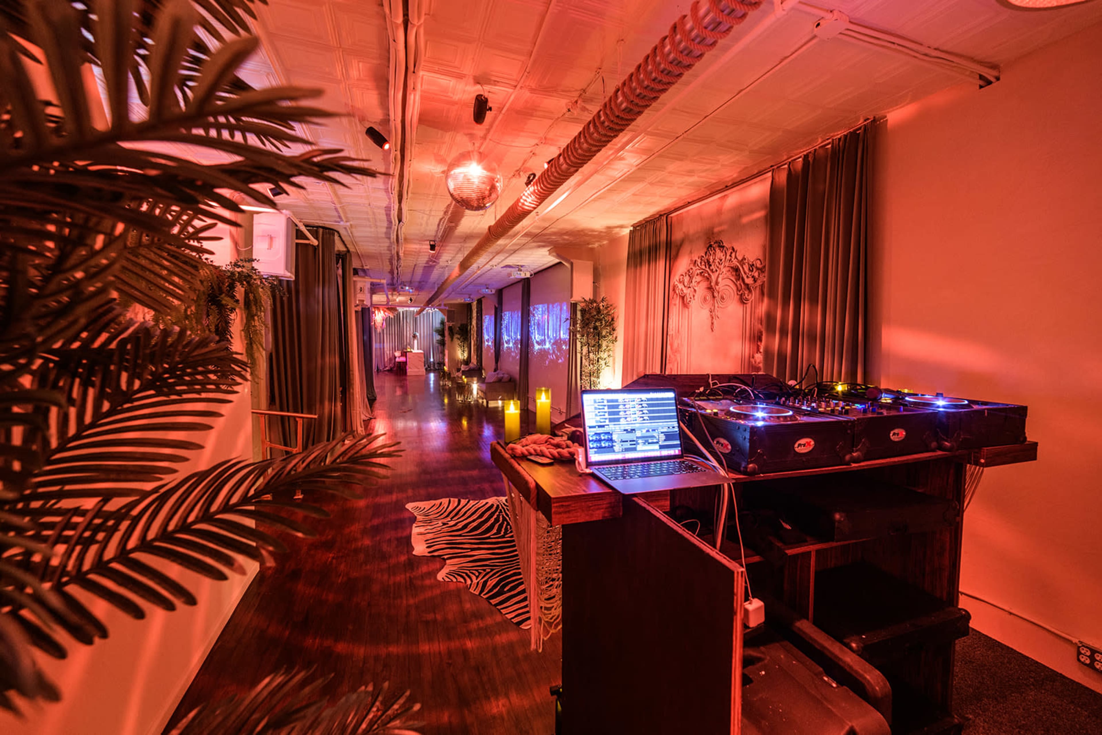 The image shows a dimly lit event space featuring a DJ booth with equipment and a laptop, surrounded by decorative elements and illuminated by ambient lighting.