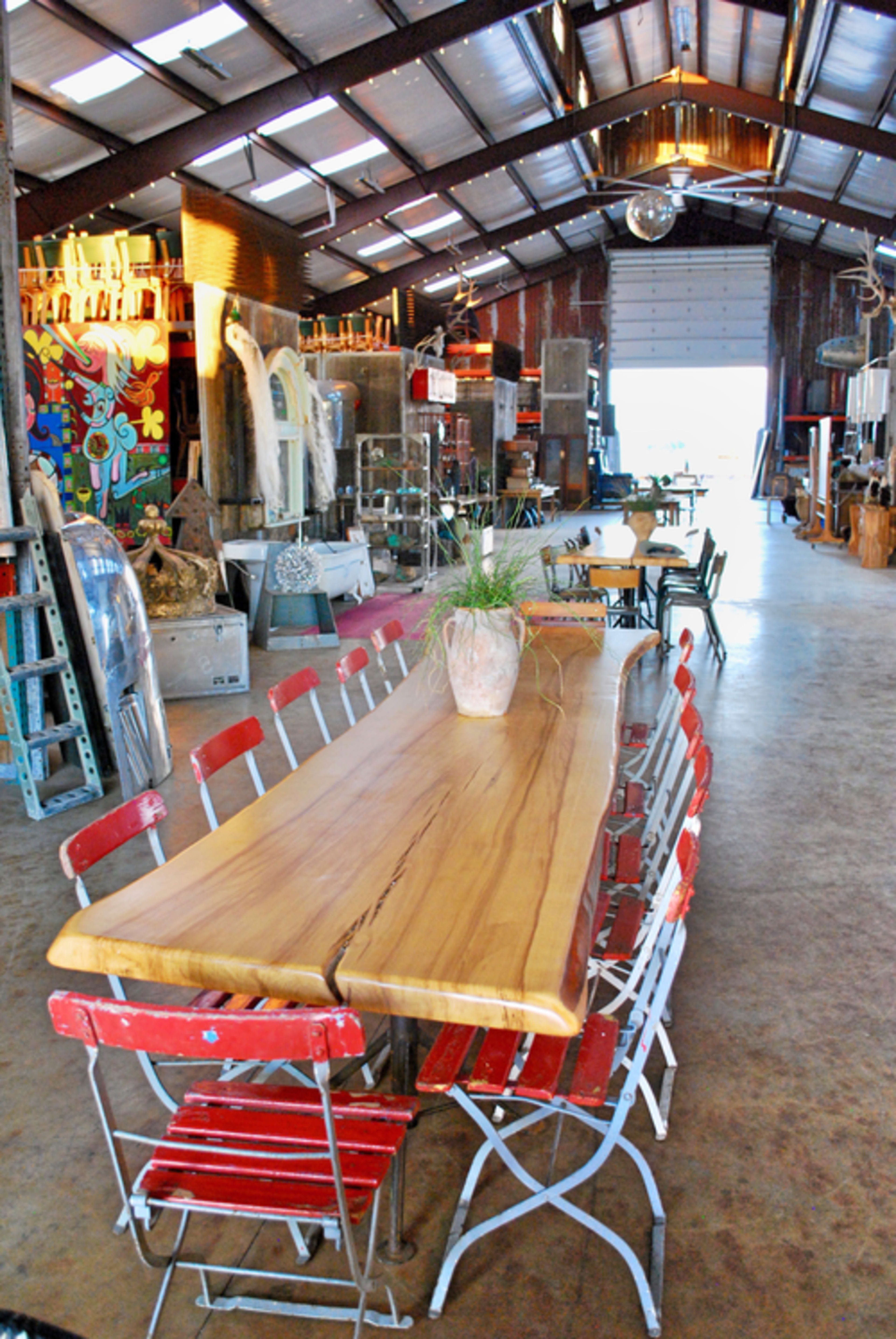 A long wooden table surrounded by red and white metal chairs is set in a spacious, well-lit barn filled with various eclectic items and artworks.
