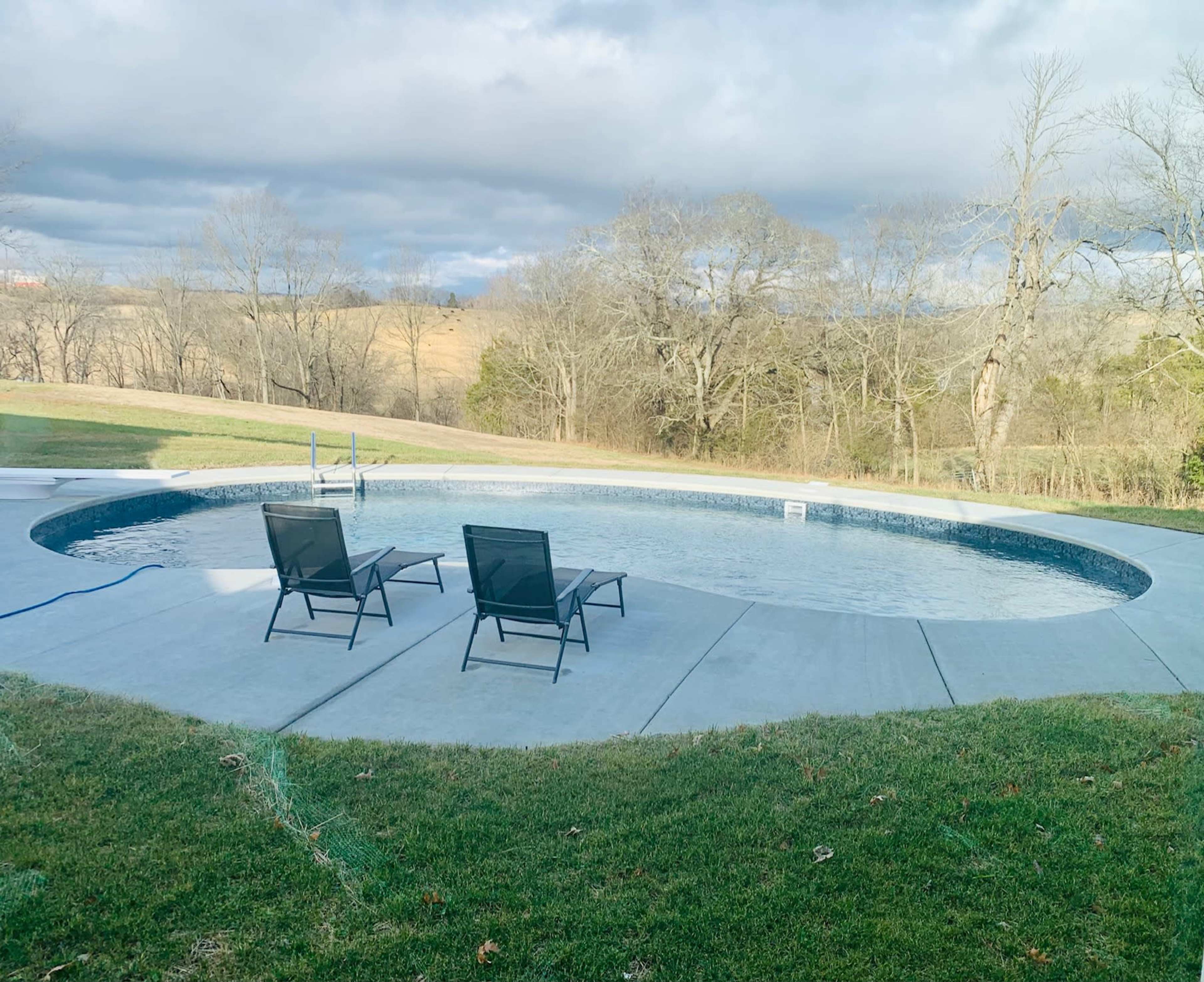 The image shows a swimming pool surrounded by a concrete deck and two black lounge chairs, with bare trees and rolling hills in the background under a cloudy sky.