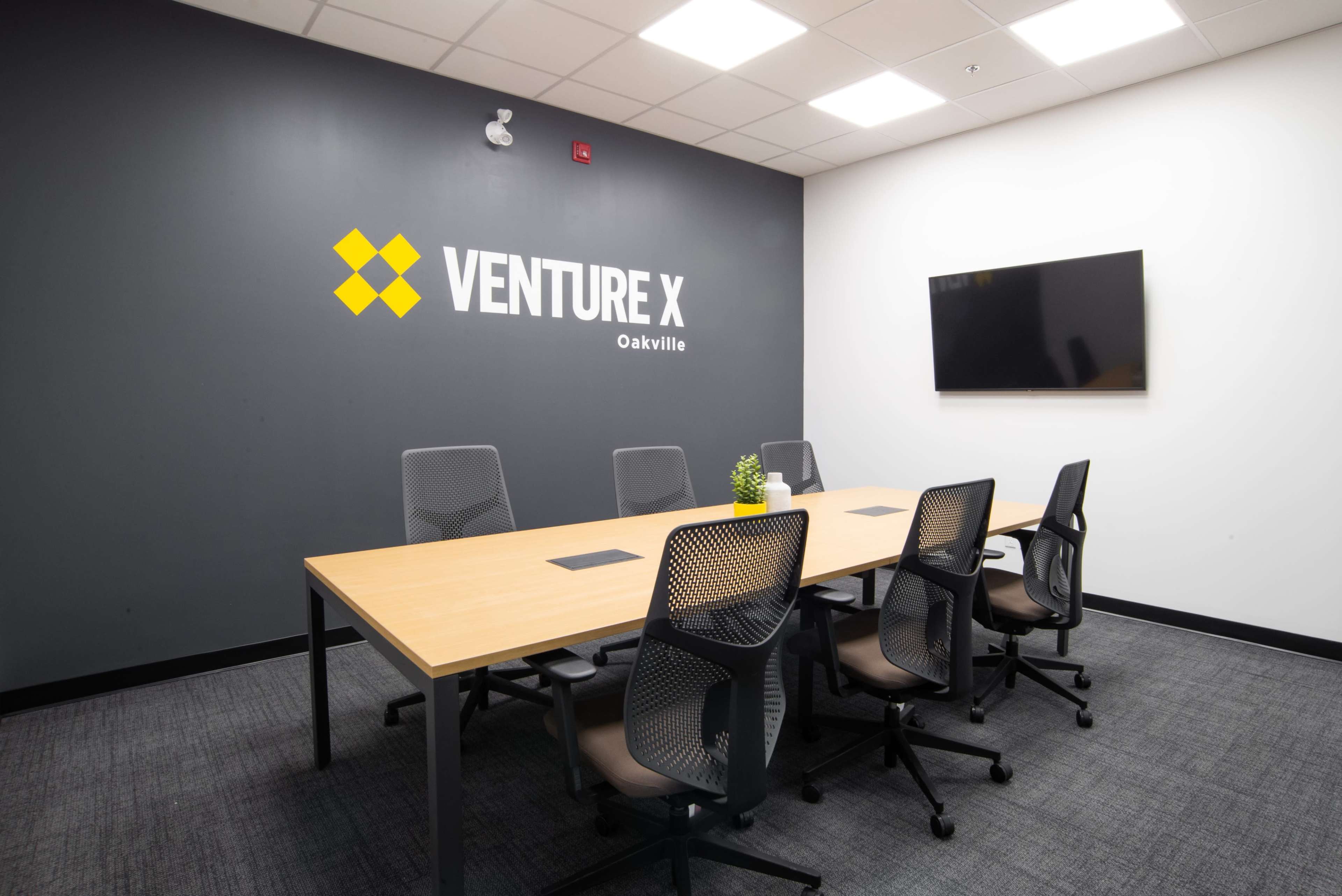 A modern conference room features a large wooden table surrounded by six ergonomic chairs, with a wall displaying the "VENTURE X Oakville" logo and a mounted television.