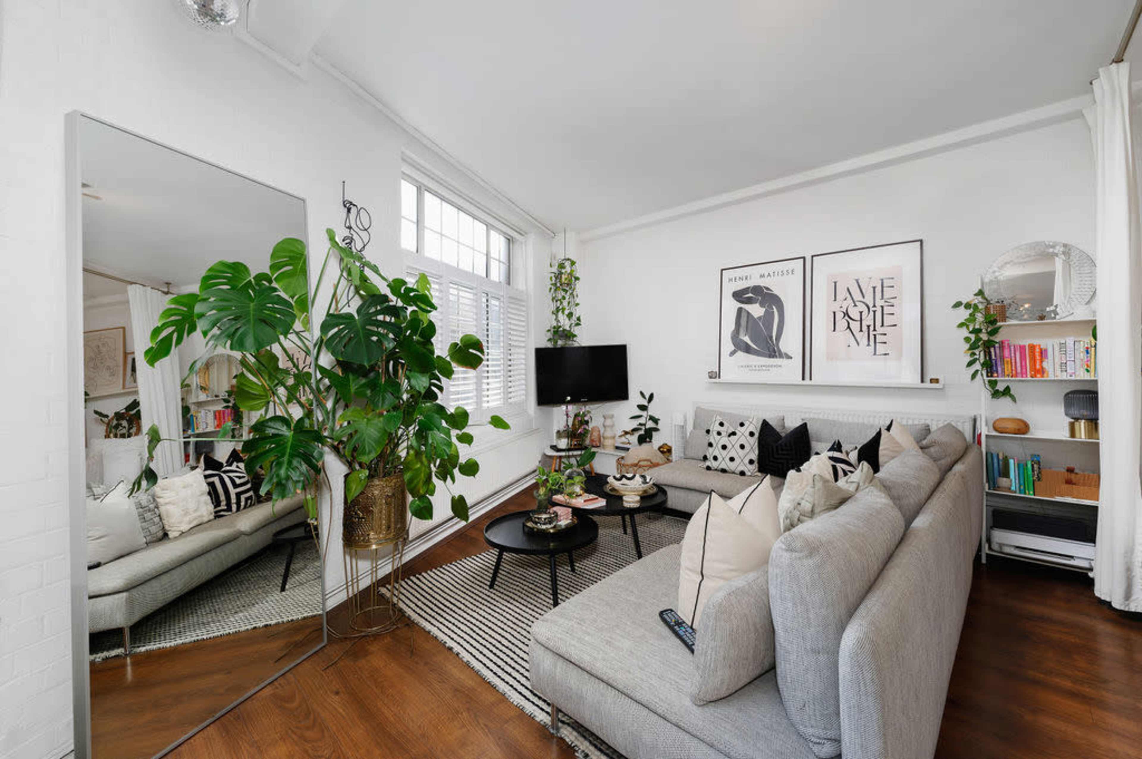 A modern living room features a sectional sofa, large mirror, various potted plants, and framed artwork on white walls.