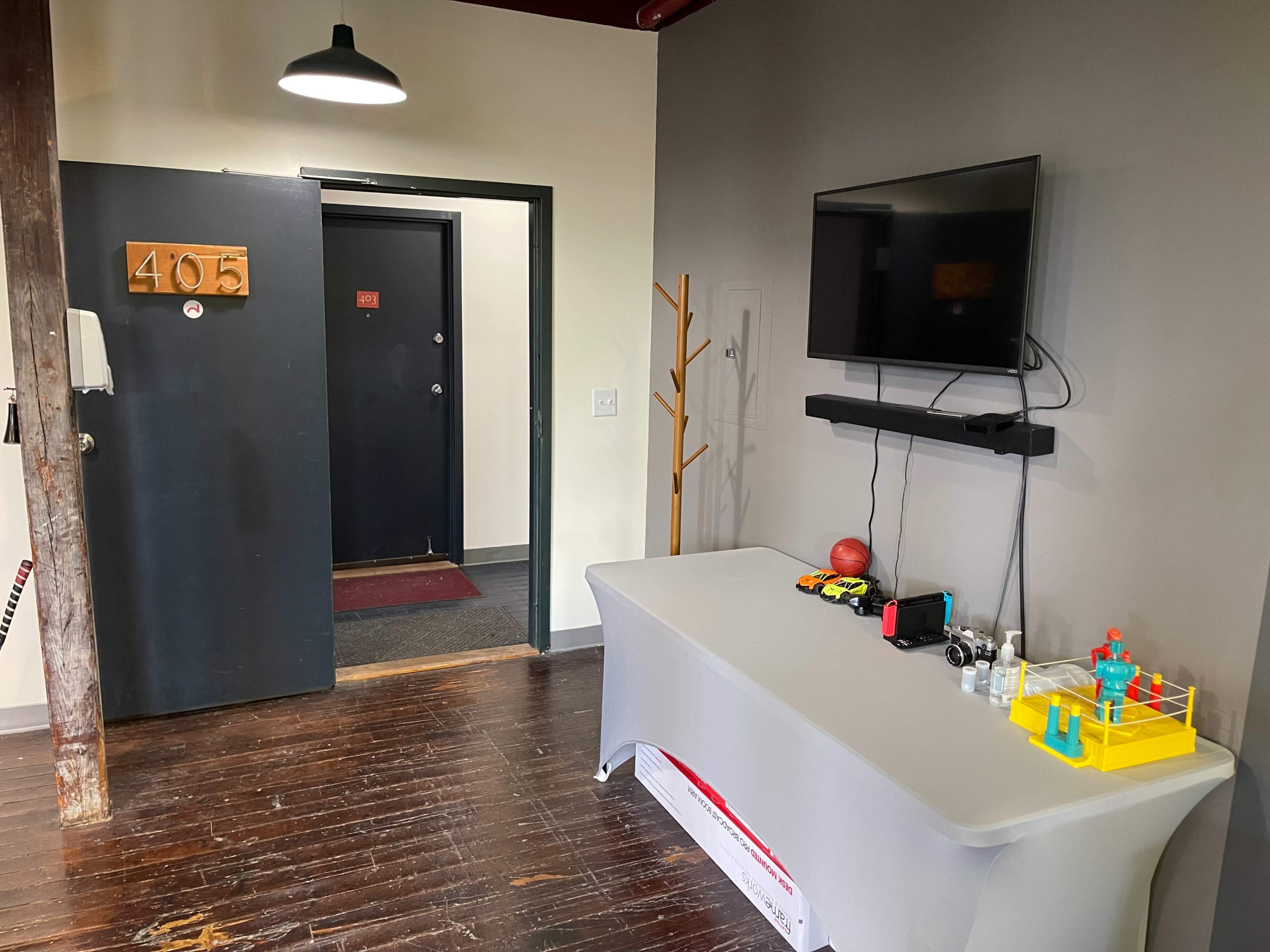 A small entryway with a door labeled "405," a coat rack, a television mounted on the wall, and a table with various items including a toy boxing ring.