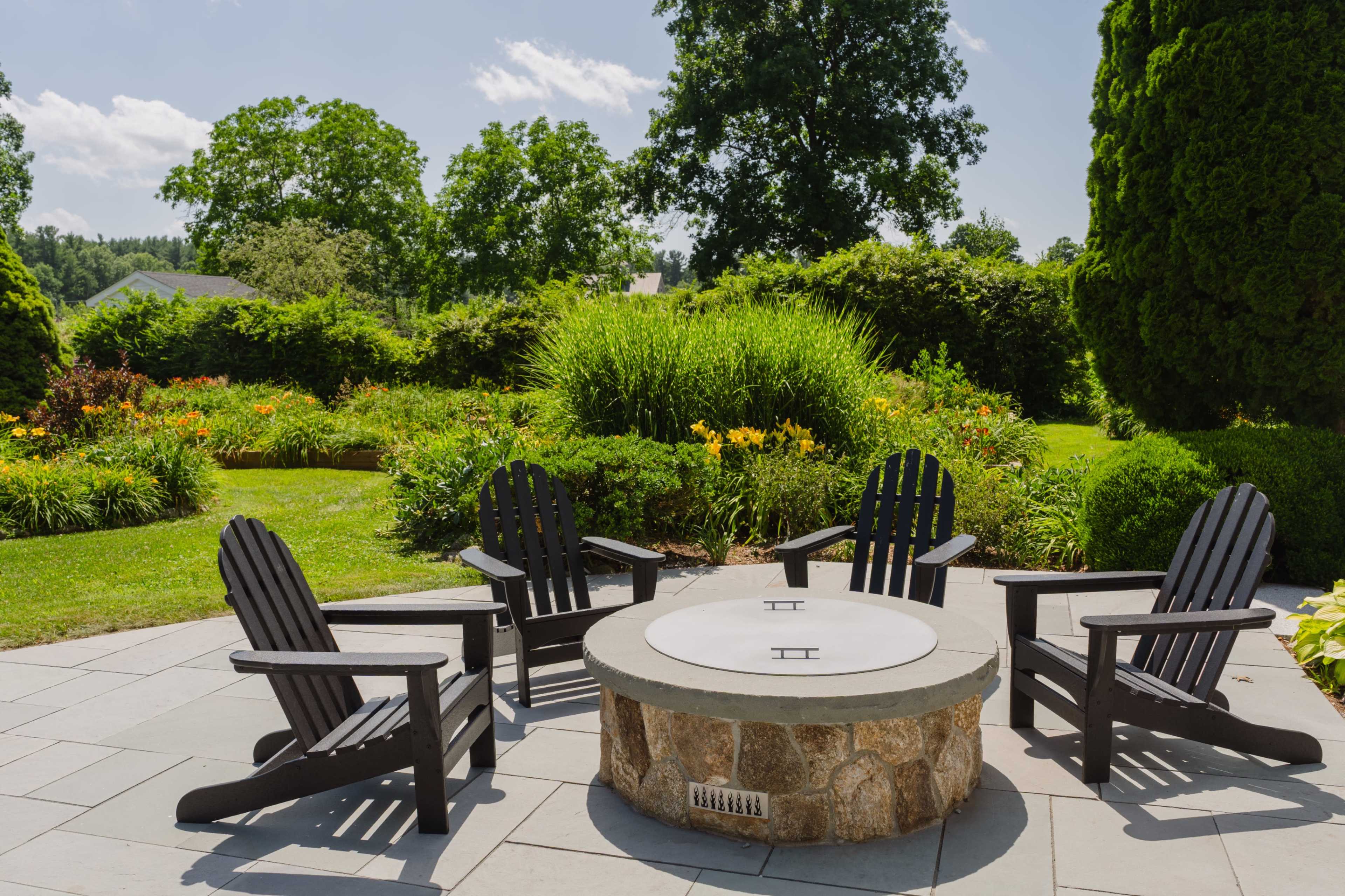 The image shows a stone fire pit surrounded by four black Adirondack chairs in a landscaped garden with greenery and flowers.