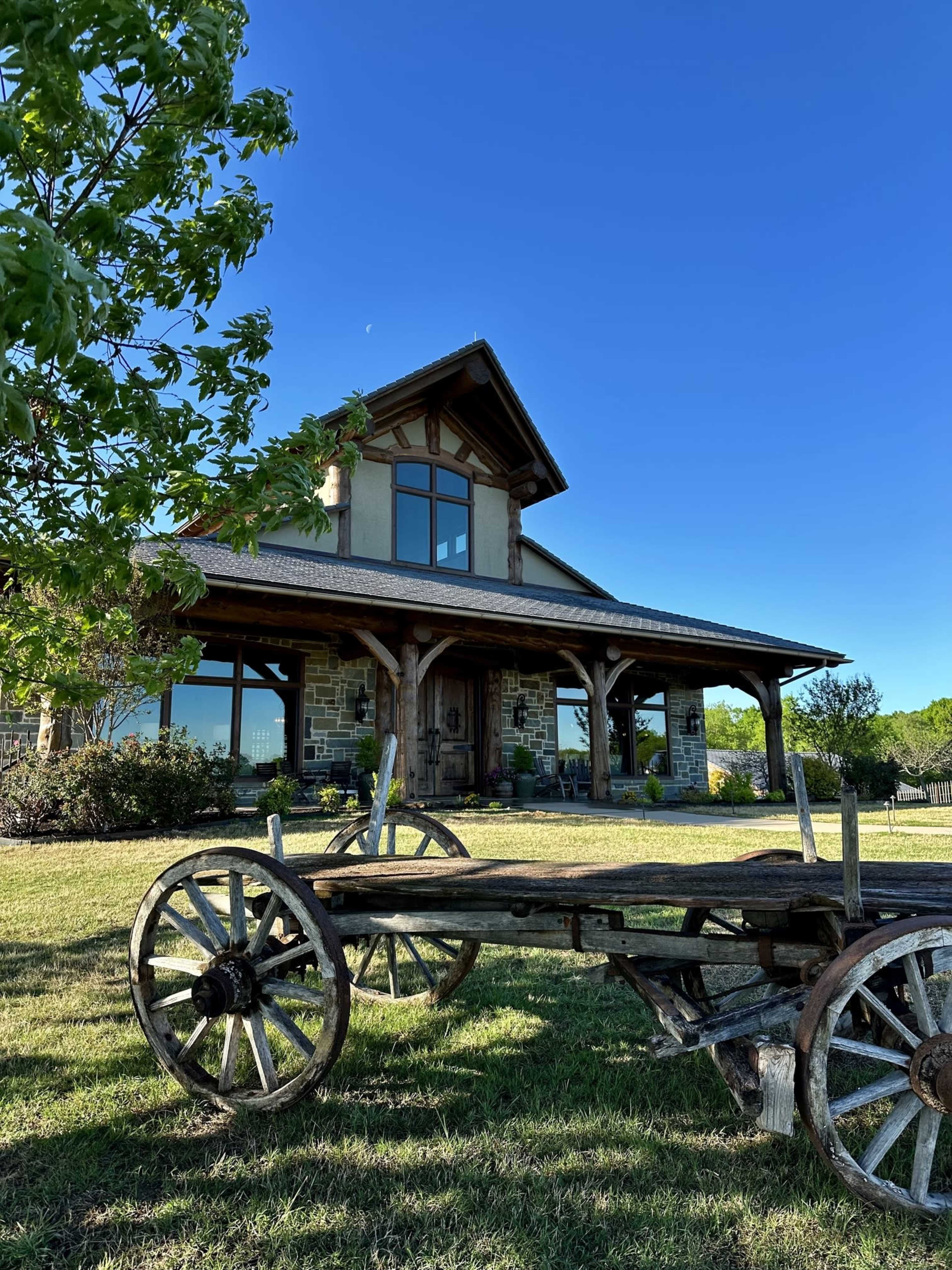 A rustic cart sits in the foreground of a stone house with large windows and a peaked roof under a clear blue sky.