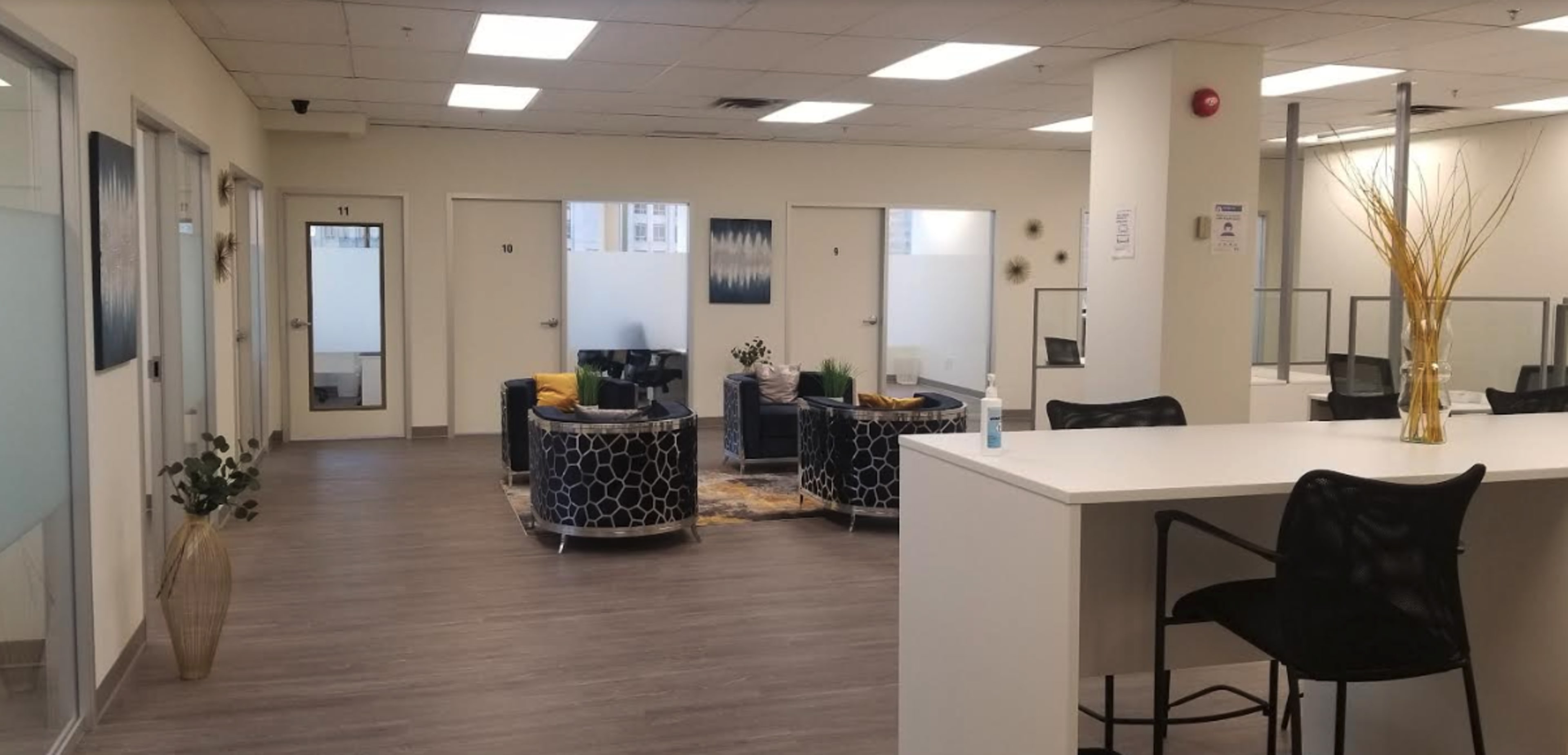 A modern waiting area with chairs, small tables, and a reception counter, featuring neutral-colored walls and a few decorative plants.