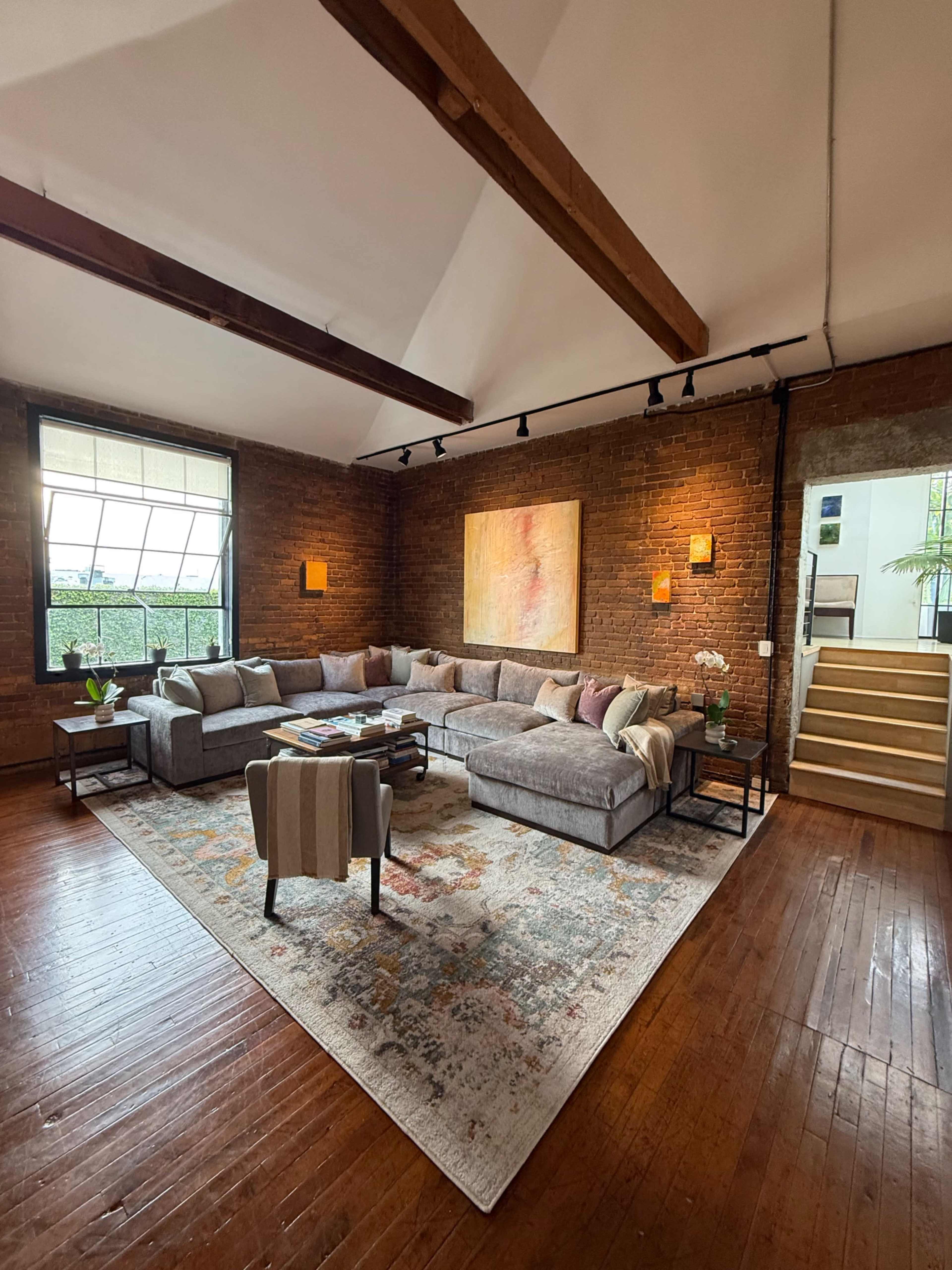 The spacious living room features a large sectional sofa, a patterned area rug, and a combination of brick walls and wooden beams.