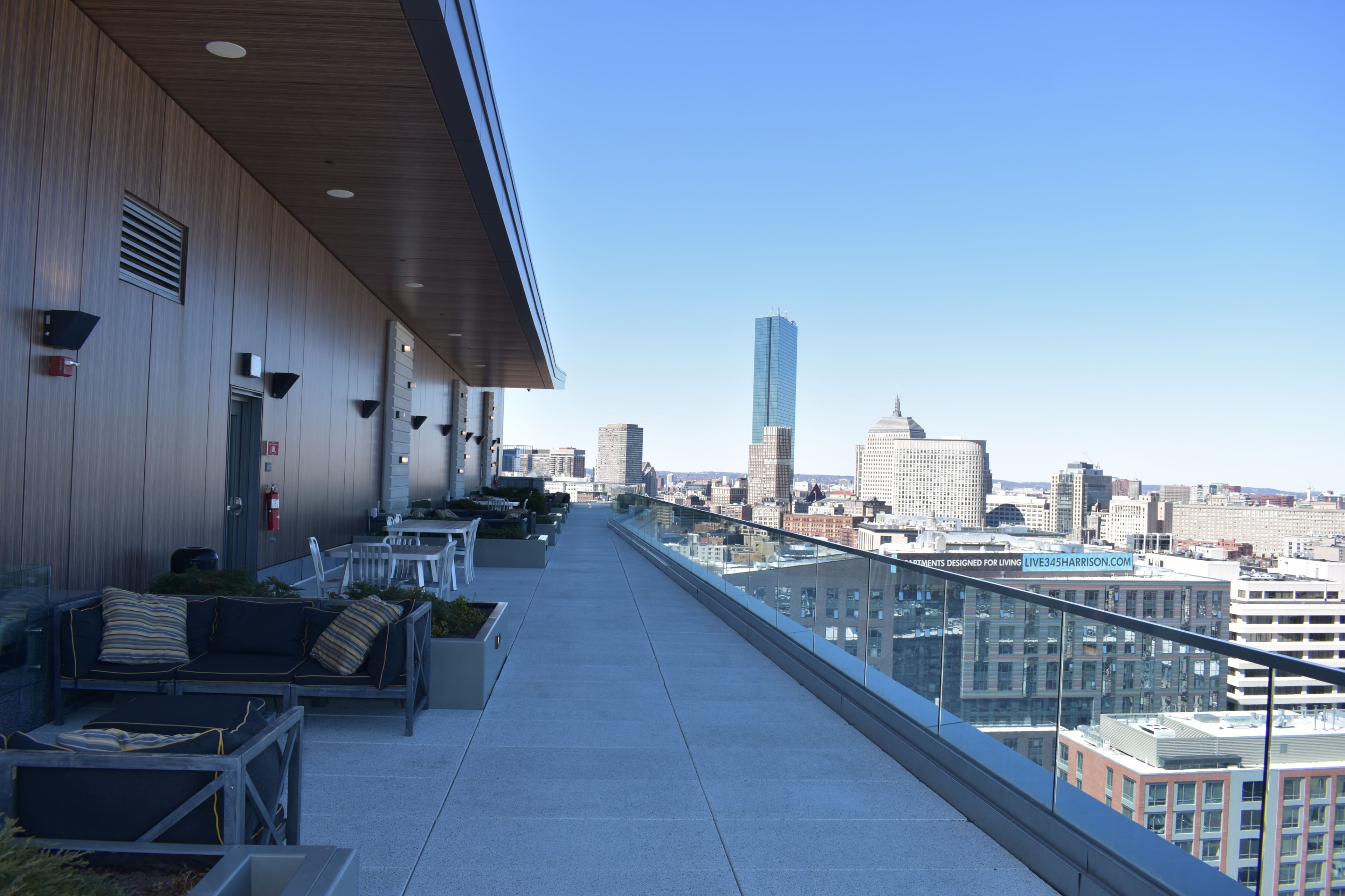 A spacious rooftop terrace with modern furniture, overlooking a city skyline featuring tall buildings and a clear blue sky.