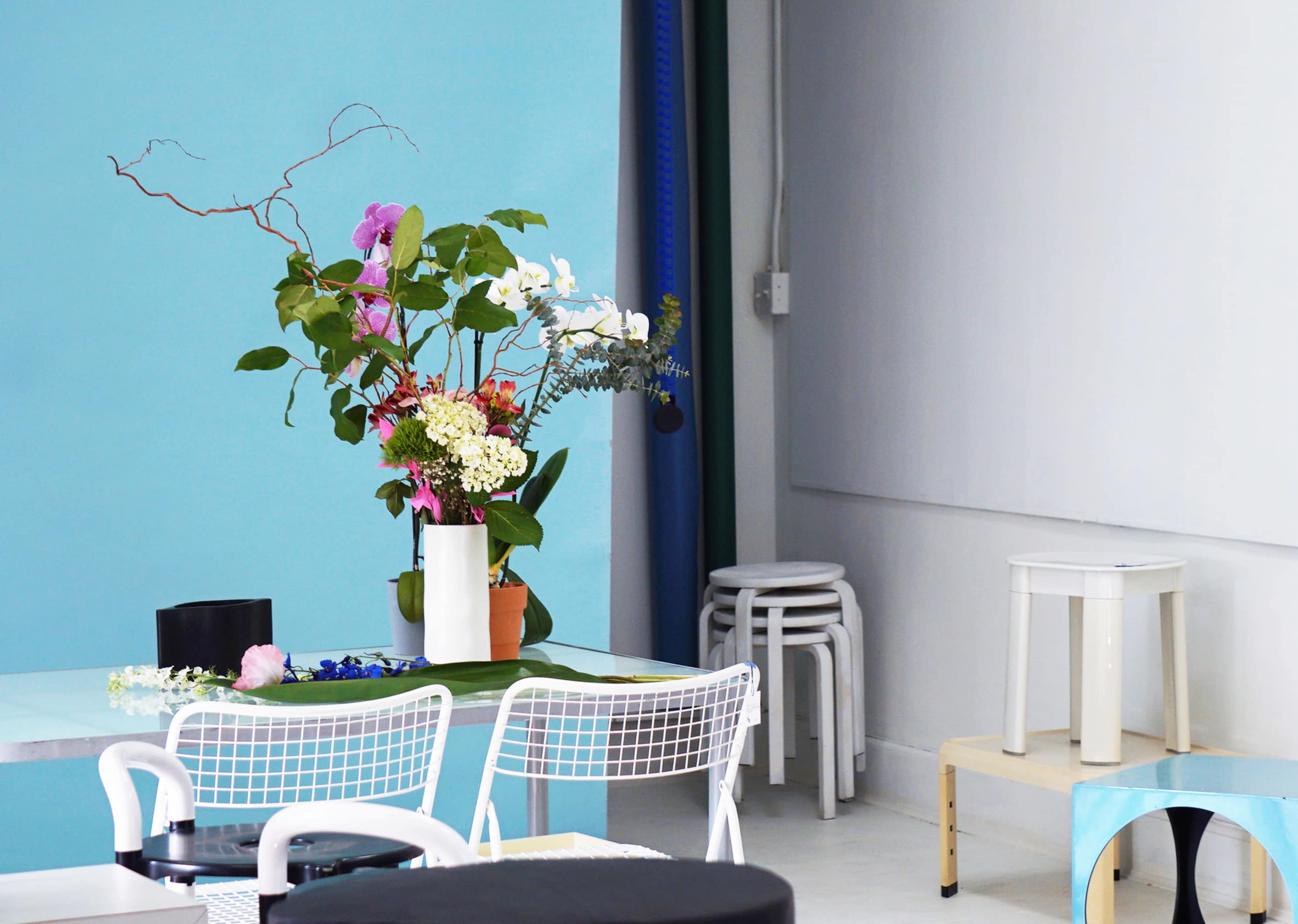 The image shows a bright, minimalist interior with a table adorned with a floral arrangement, surrounded by modern chairs and stools against a backdrop of blue and white walls.