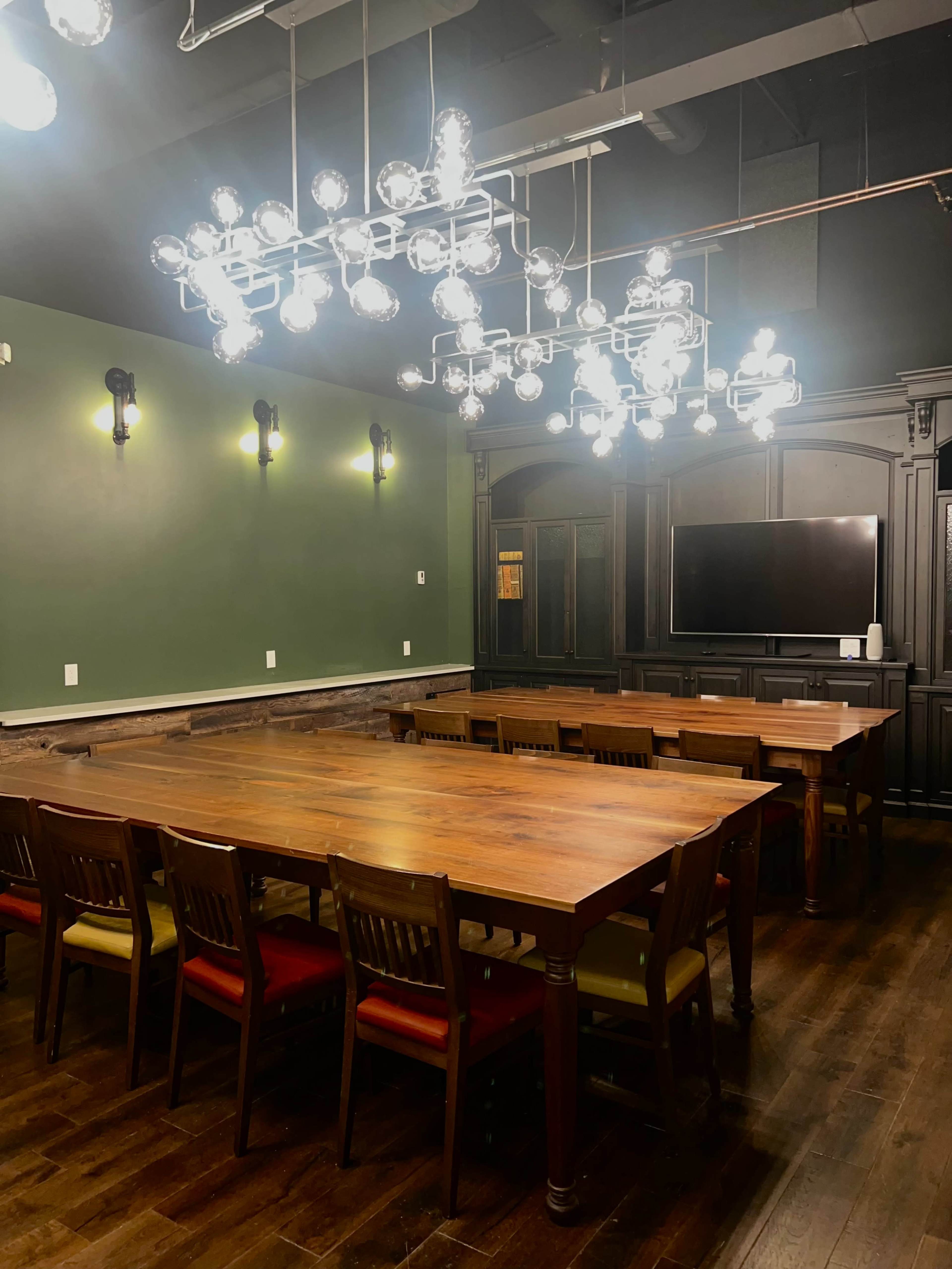 The image shows a well-lit conference room featuring a long wooden table surrounded by chairs, with a large wall-mounted television and a modern chandelier overhead.