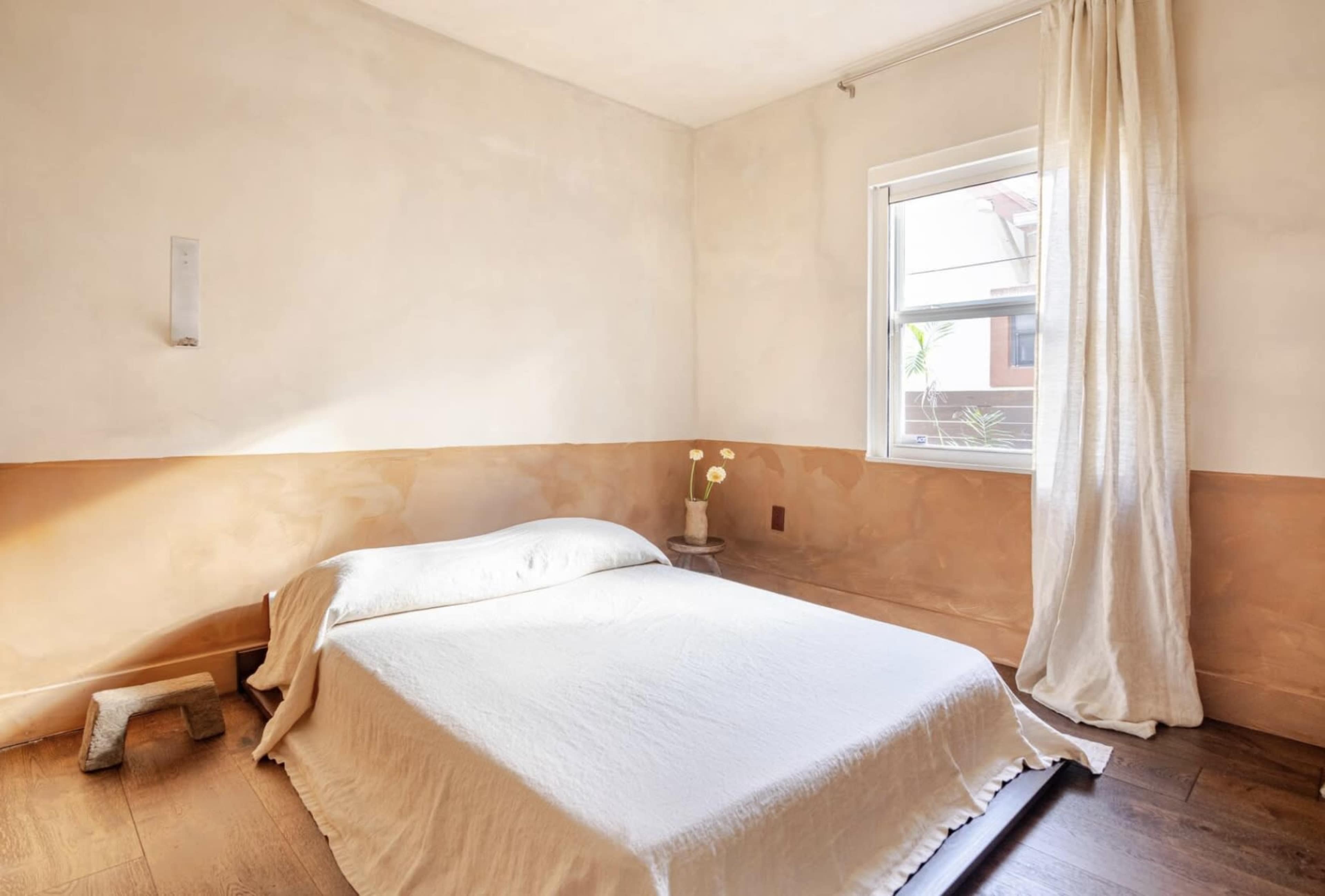 The image shows a minimalist bedroom featuring a bed with a white blanket, wooden flooring, and a window with sheer curtains.