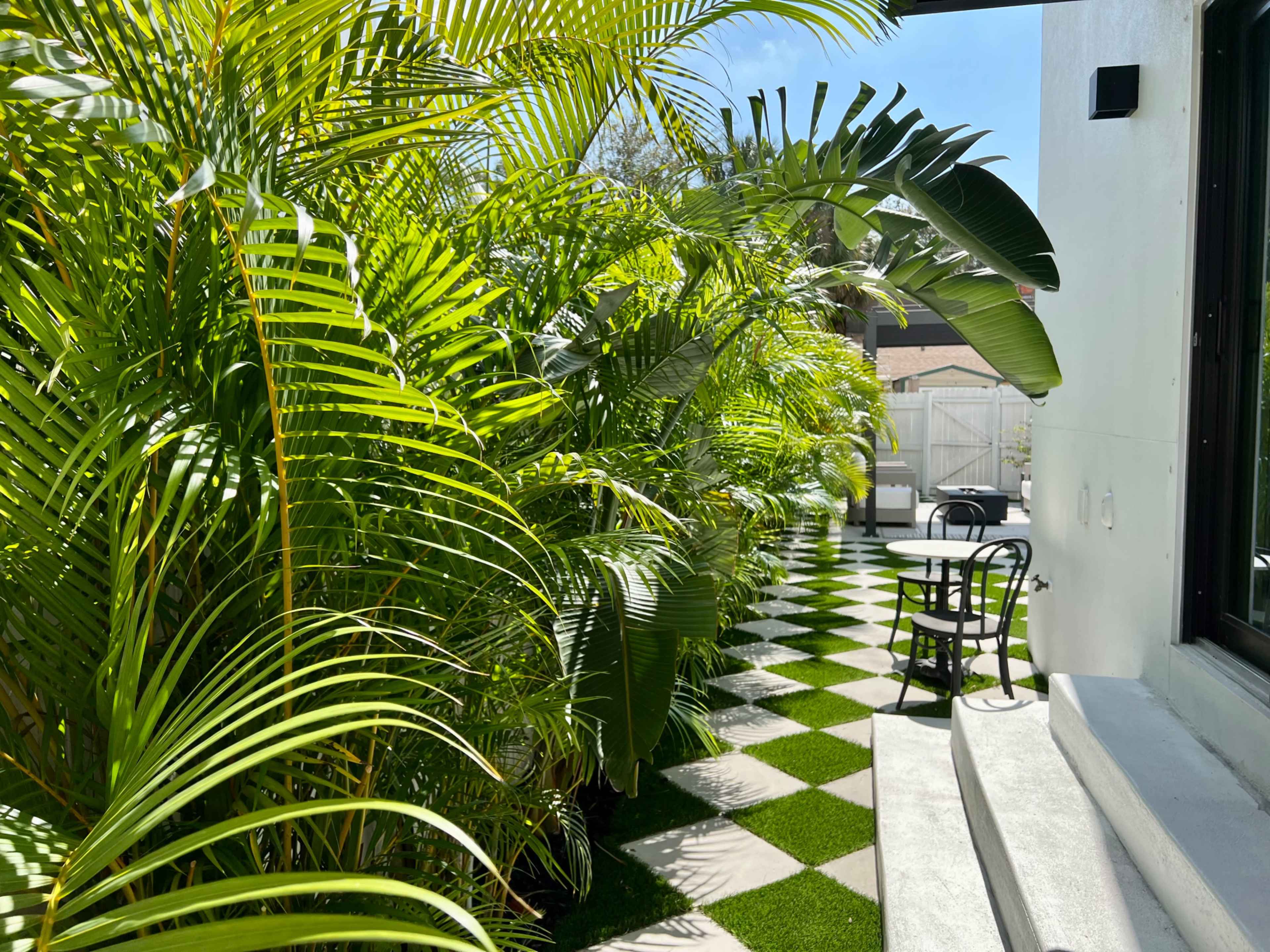 A path lined with lush green palms leads to a small outdoor seating area with a black-and-white checkered floor.