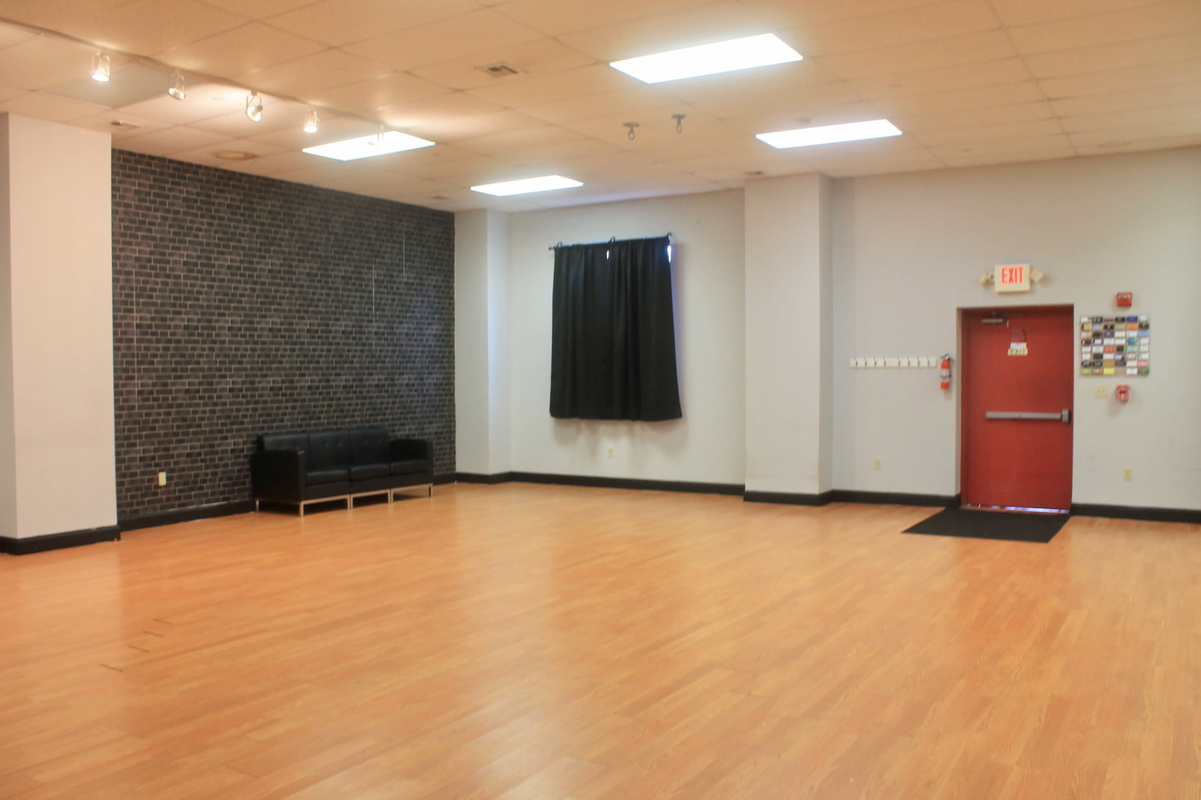 A spacious room with wooden flooring, a black sofa against a textured wall, a window covered by a black curtain, and a red exit door.