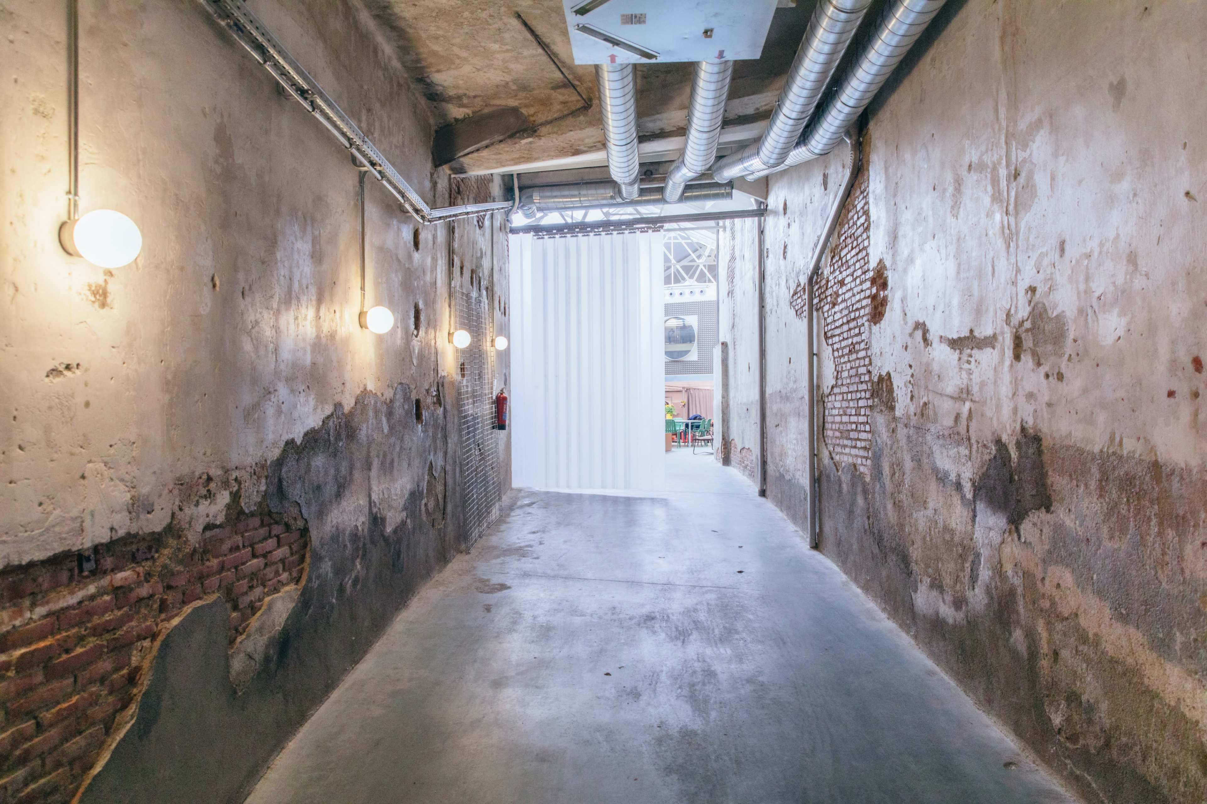The image shows a long, narrow corridor with exposed brick walls, industrial-style lighting, and a white partition at the end.