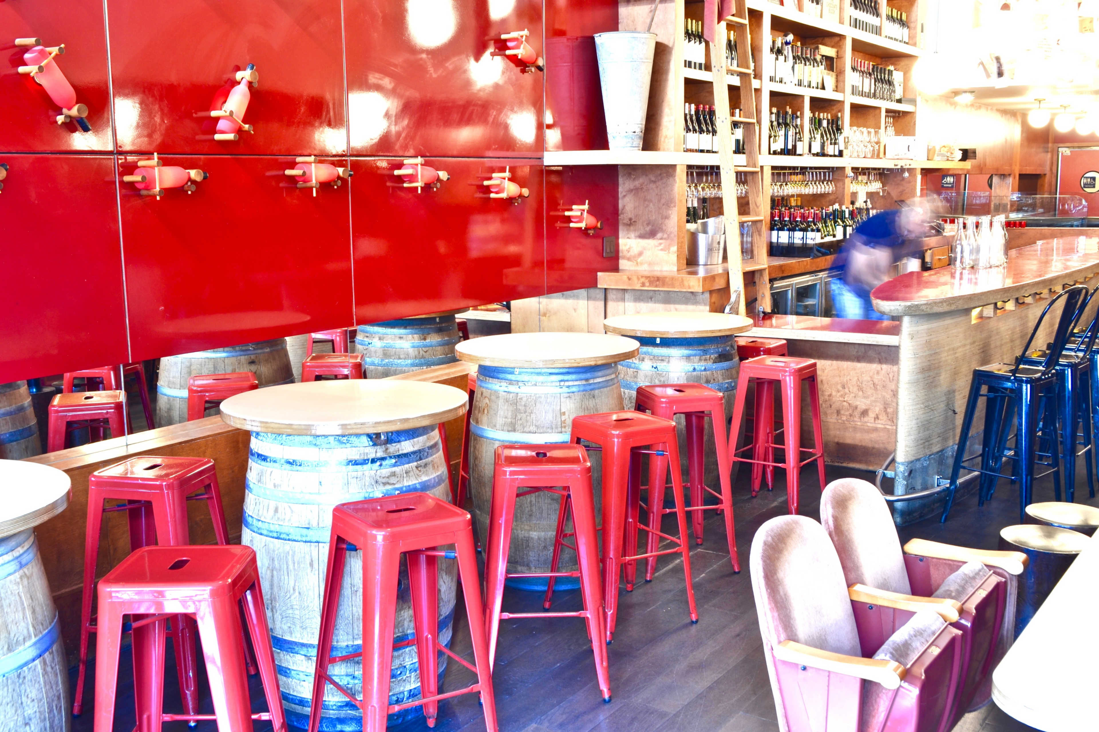 A bar interior featuring red metal stools and wooden barrels as tables, with shelves of wine bottles in the background.