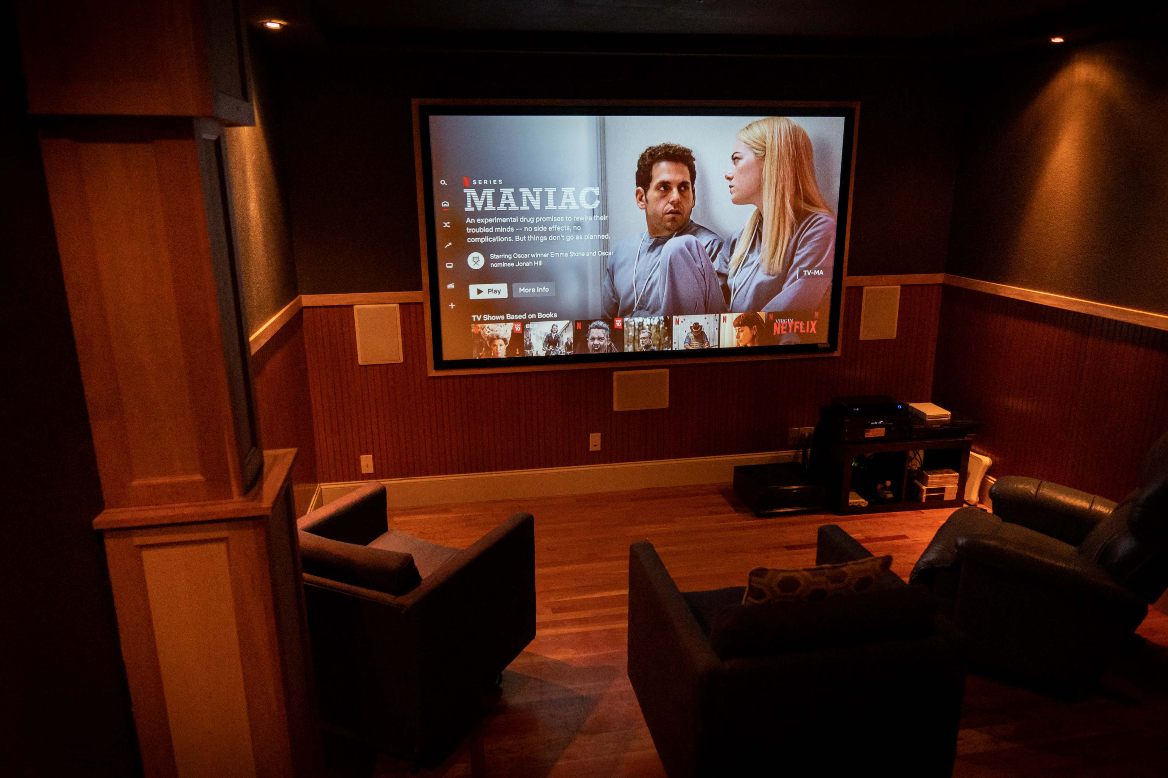 A home theater features a large screen displaying the Netflix series "Maniac" with two characters, and two comfortable chairs are positioned in front of it.