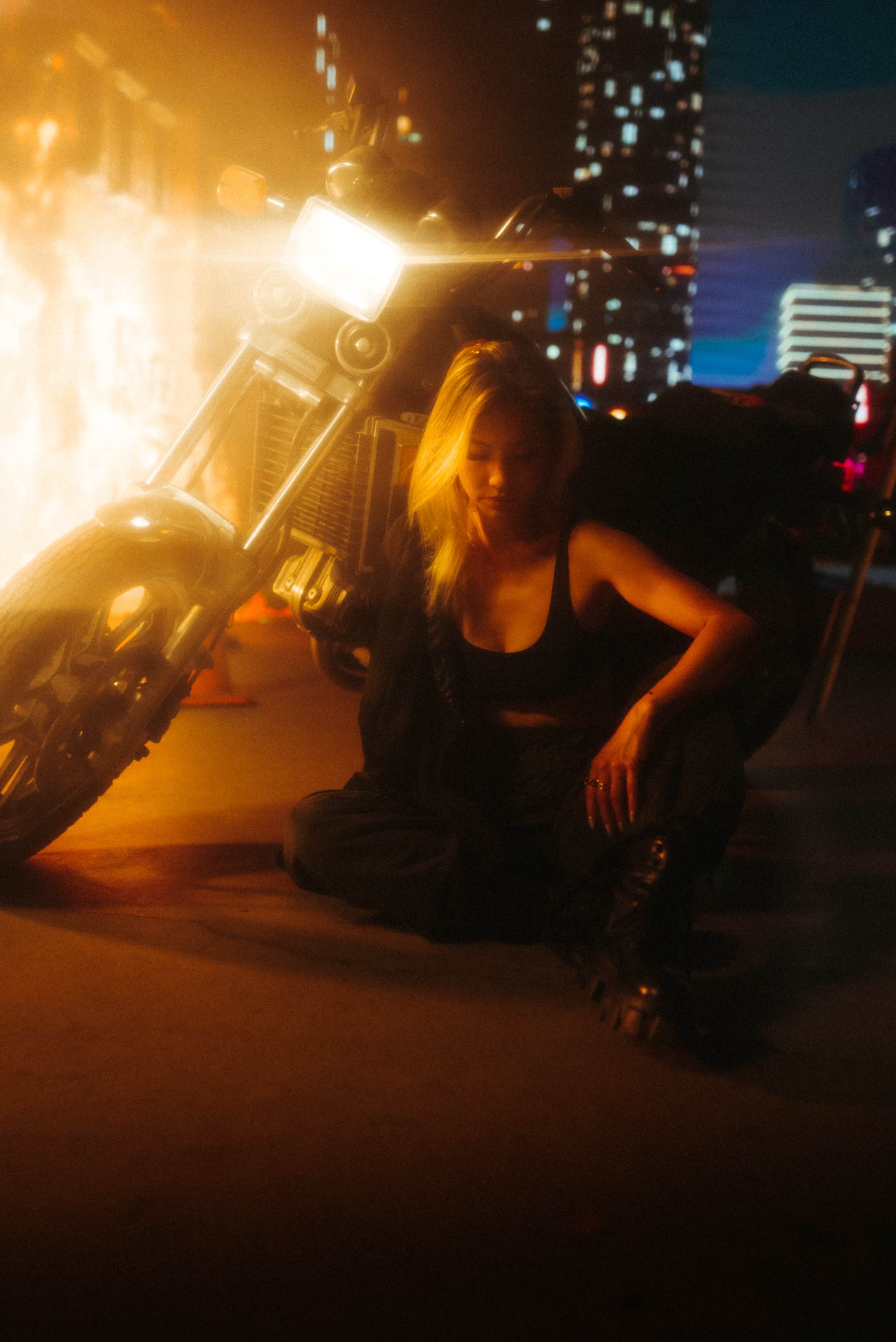 A woman with blonde hair sits on the ground next to a motorcycle amid a brightly lit urban backdrop.