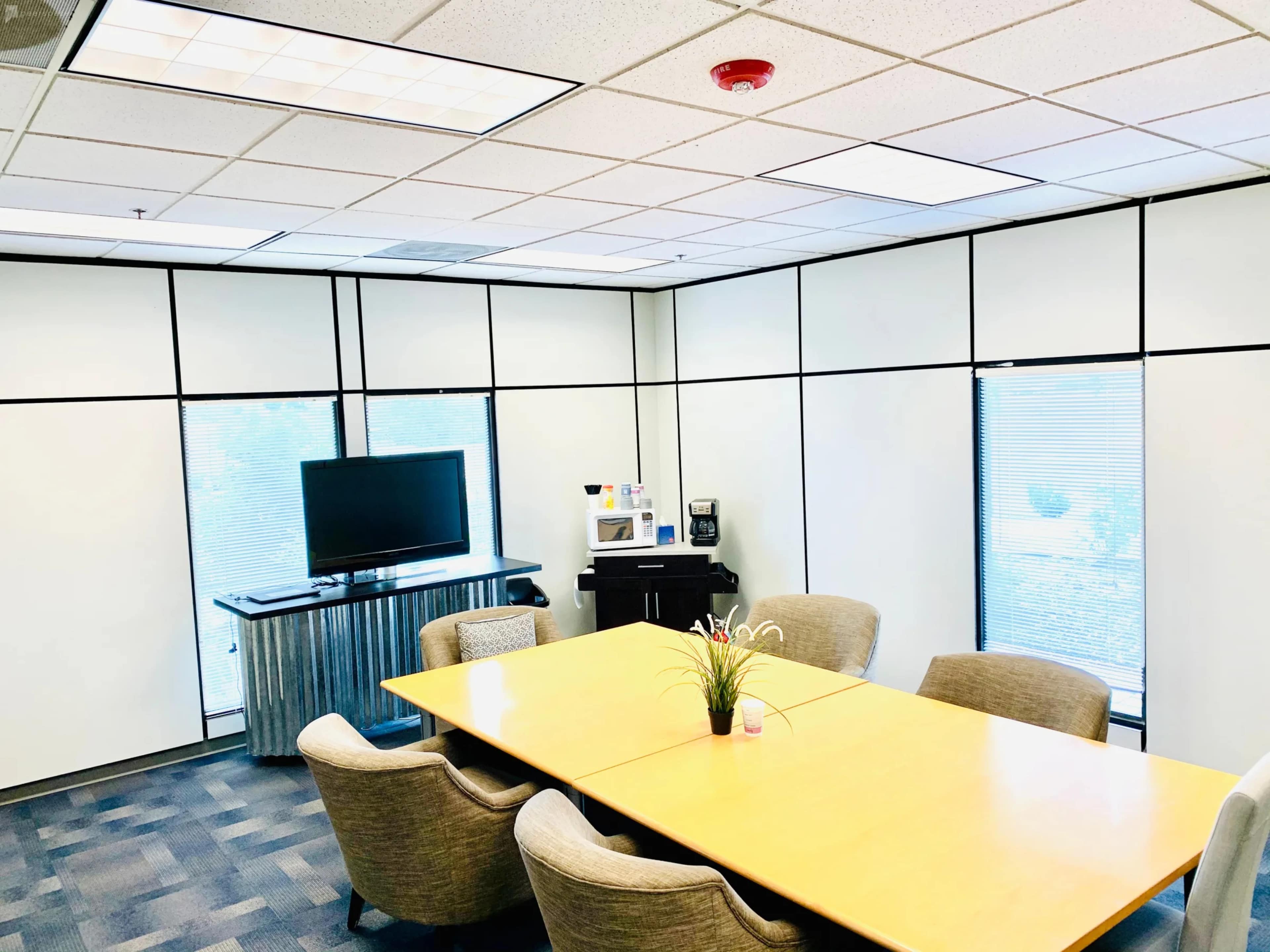 A conference room features a large wooden table surrounded by beige chairs, a television mounted on the wall, and a kitchenette area with a coffee machine.