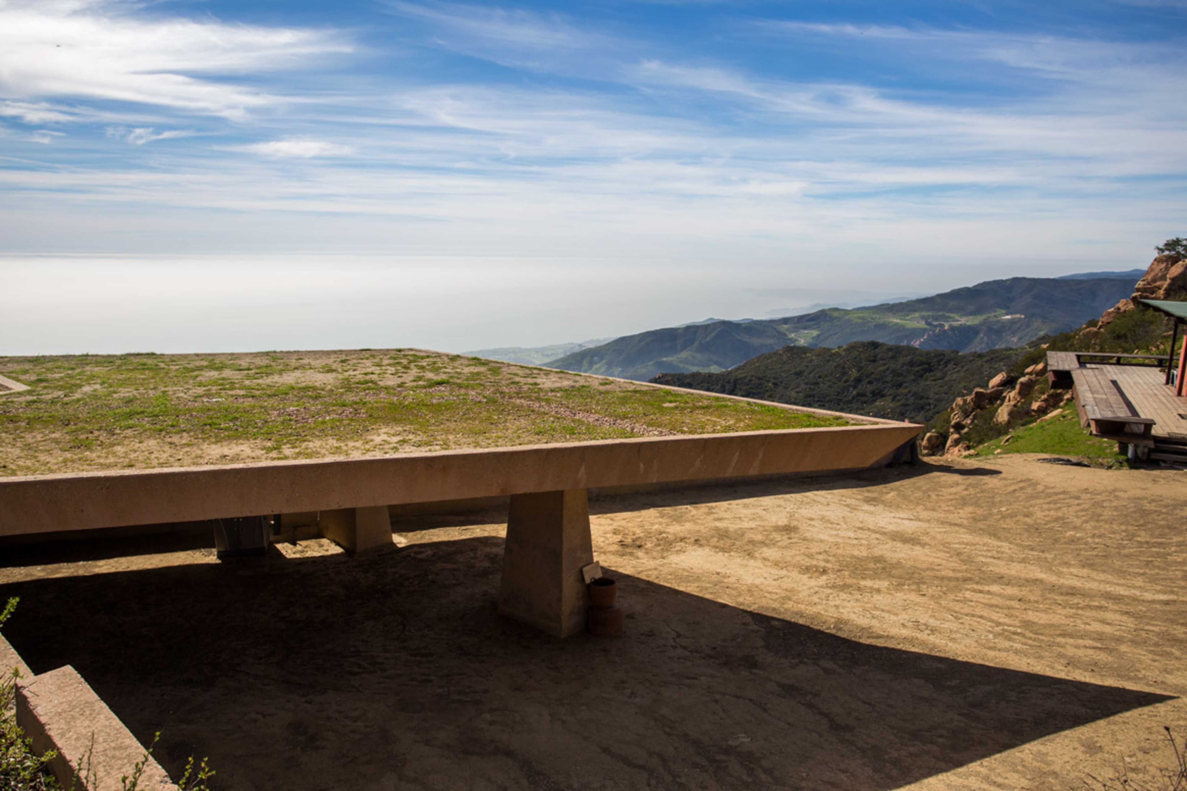 A flat, grassy rooftop structure extends over a rocky hillside, overlooking a vast landscape and the sea beyond.